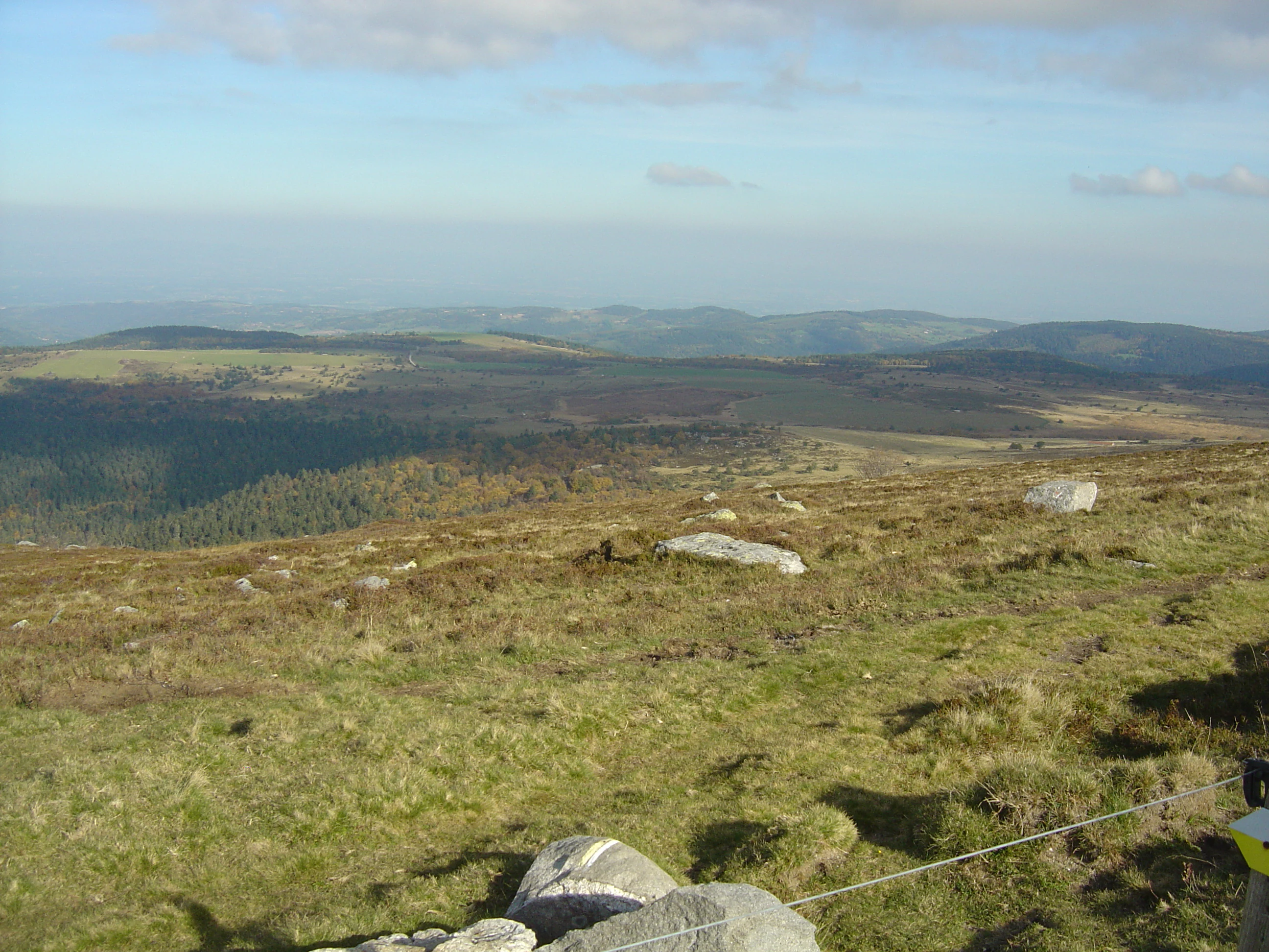 A wide, high-angle view of grassy, rolling highlands with scattered rocks and distant hills under a partly cloudy sky.