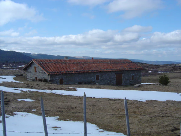 A long, single story stone building with a red tiled roof, situated in a high altitude field with patches of melting snow.