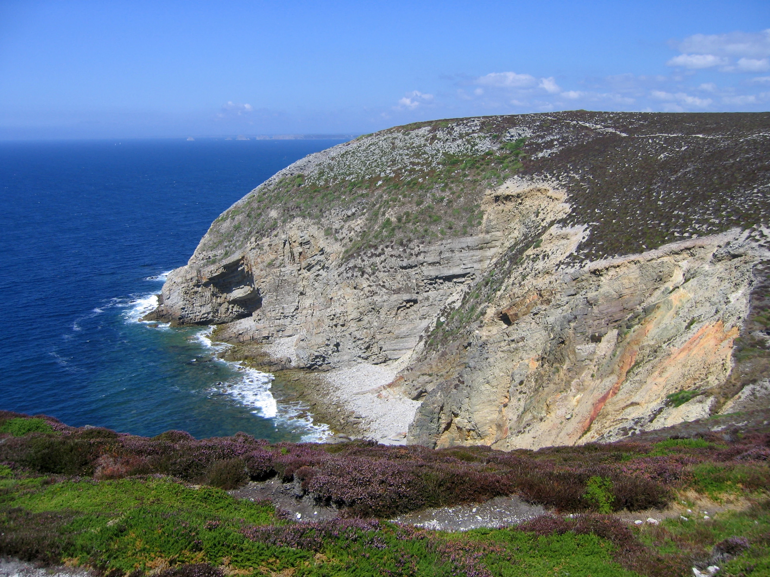 A wide view of a large, rocky headland protruding into the deep blue ocean, with exposed geological cliff faces and low-lying coastal vegetation in the foreground.