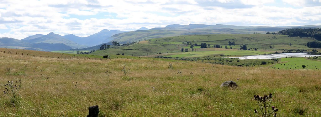 A wide, panoramic view of a grassy highland plateau with gentle hills and distant mountains under a cloudy sky.