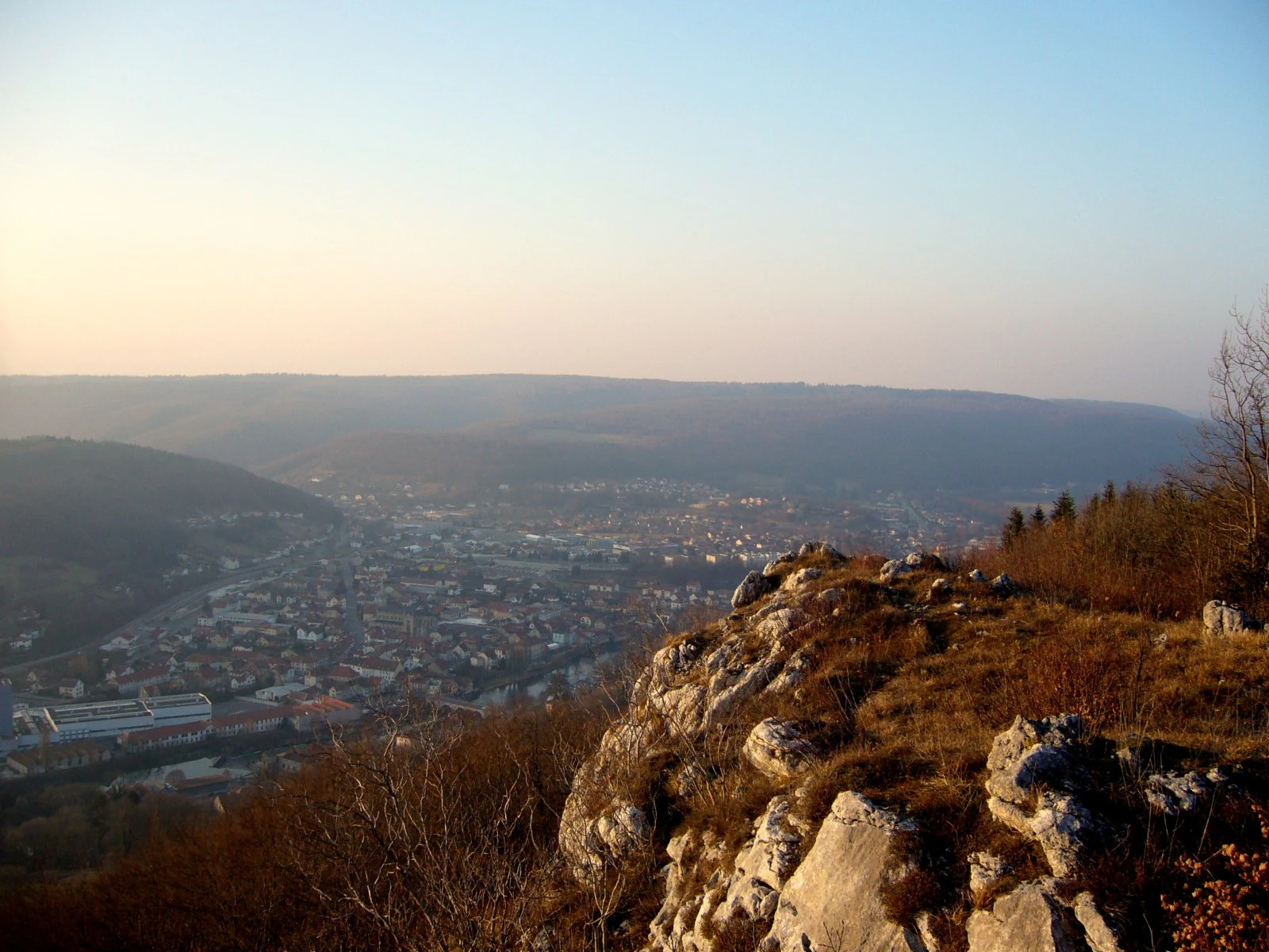 A high vantage point looking down over a town nestled in a valley, with rugged limestone rock formations in the foreground under a hazy sky.