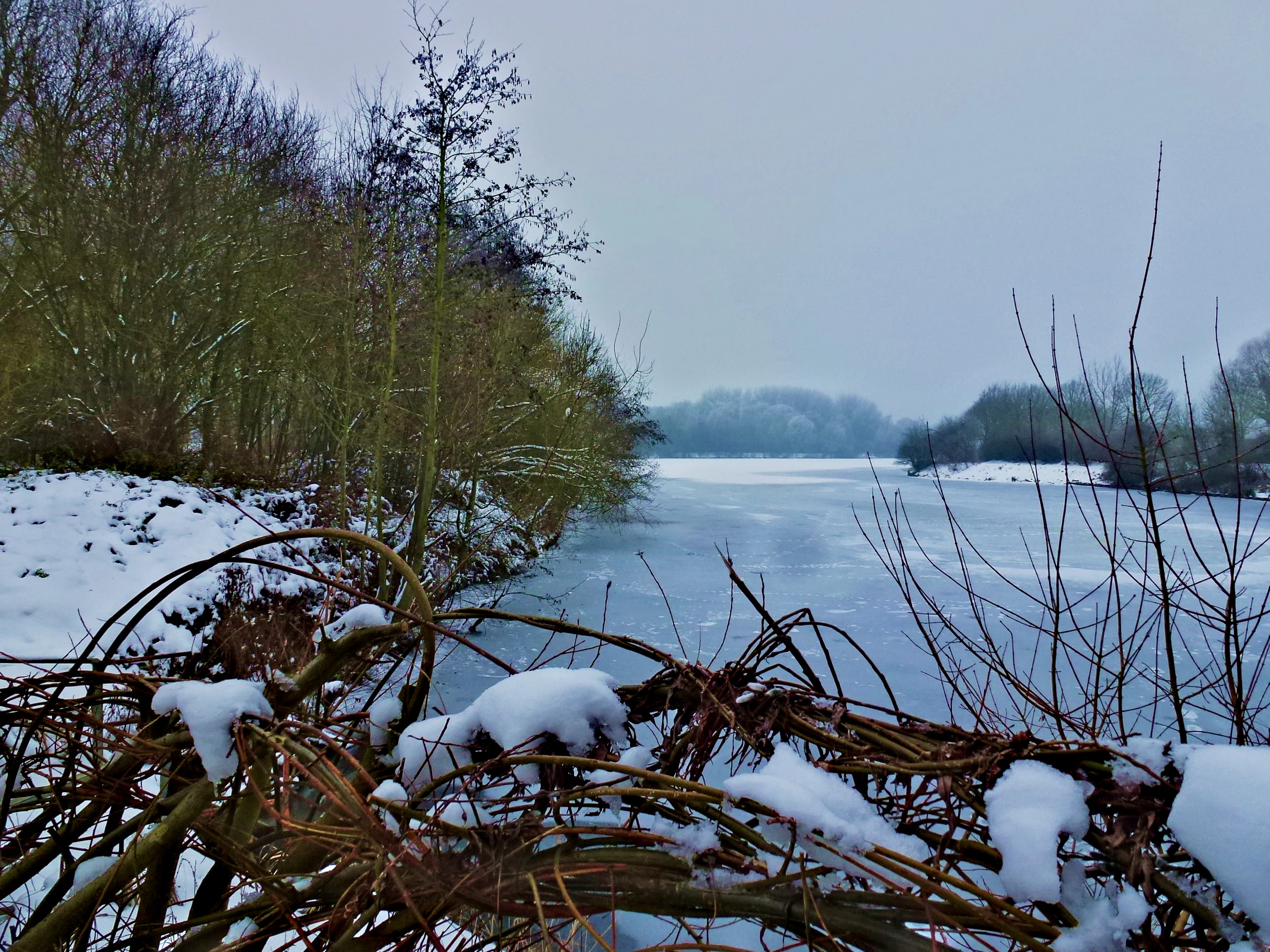 A frozen lake surrounded by snow covered banks and winter trees under a grey, overcast sky.