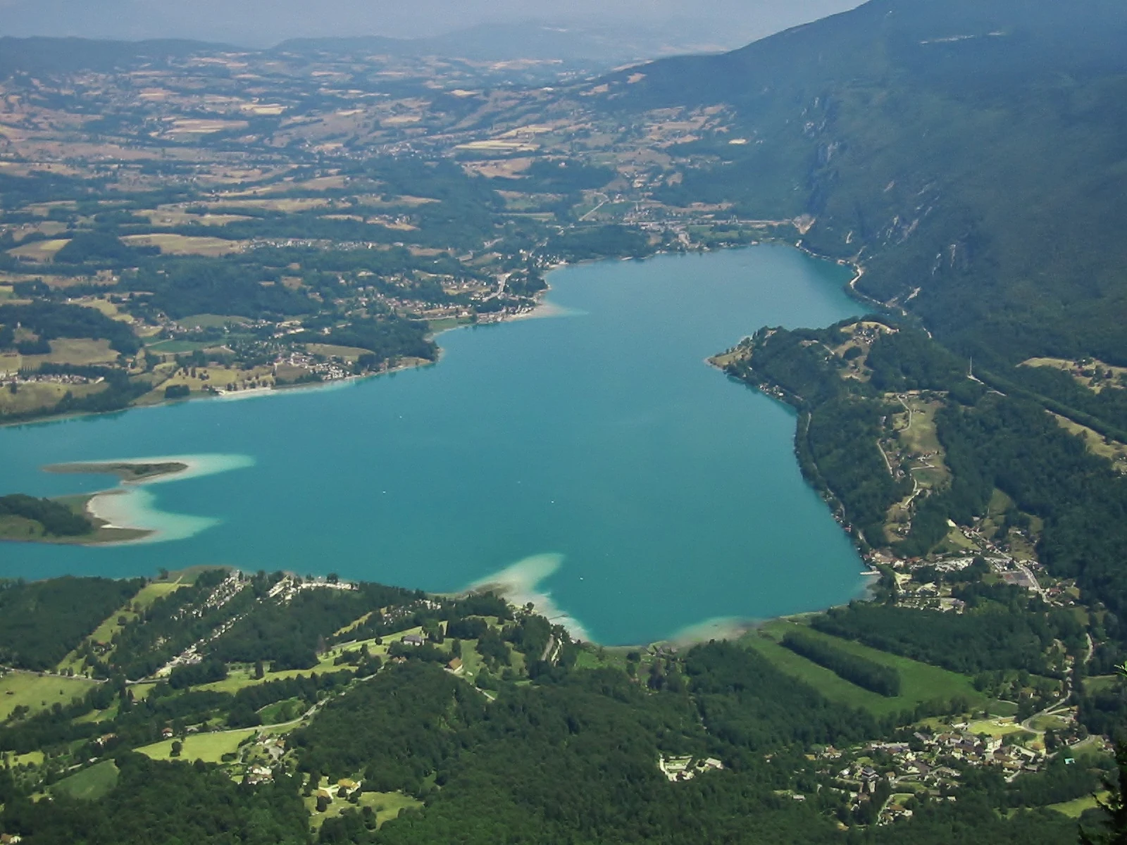 A wide aerial view of the turquoise-colored Lake Aiguebelette surrounded by forested mountains and scattered rural development.