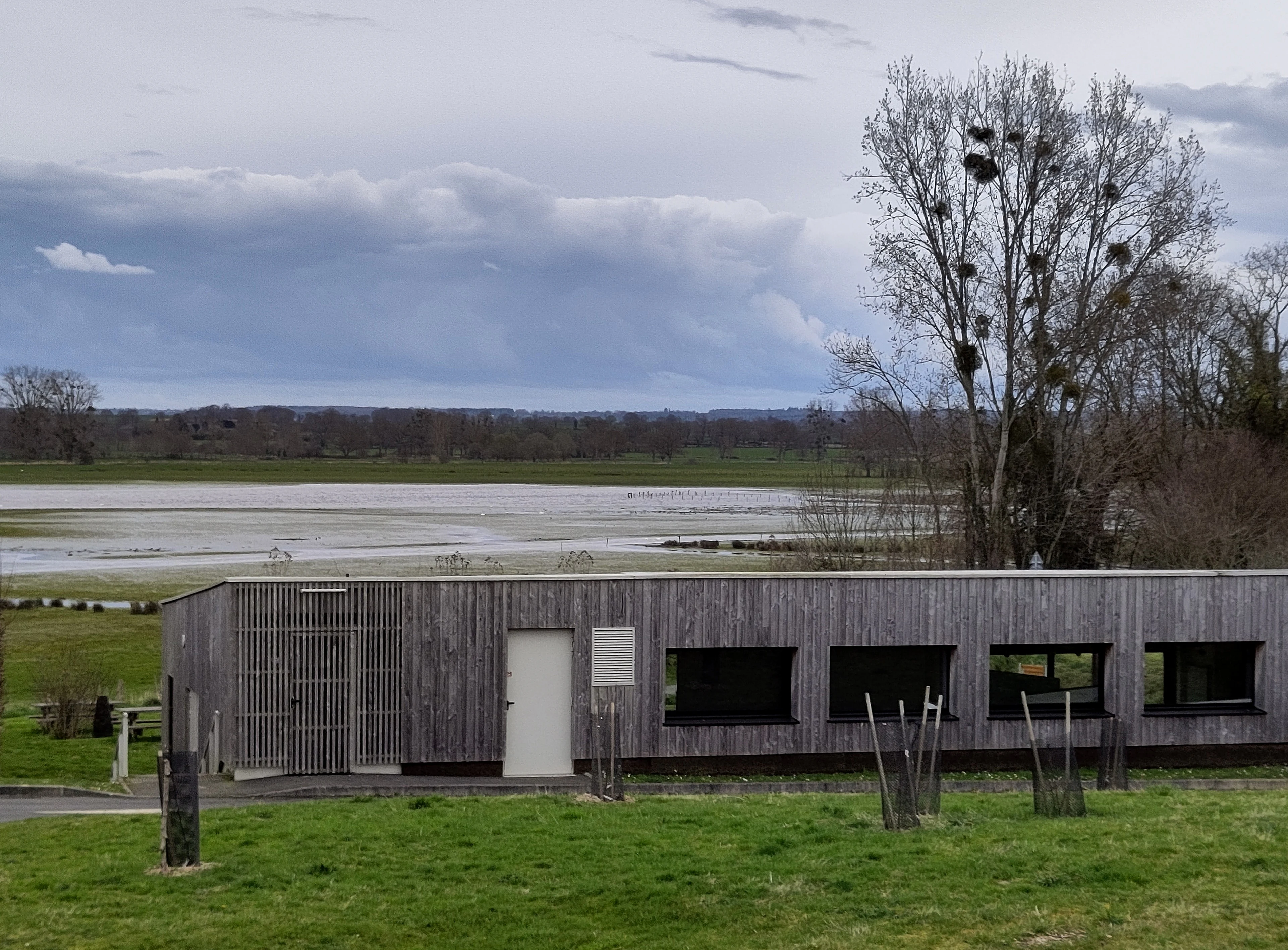 A modern, single-story building with grey vertical wood cladding sits in a green field, overlooking a wetland area under a cloudy sky.