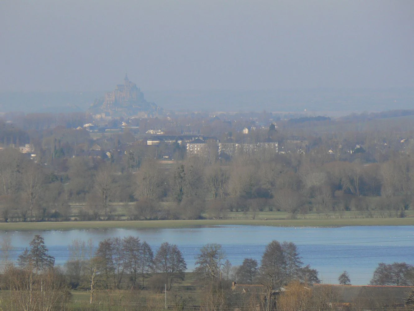 A wide, hazy, high-angle landscape shot showing a body of water in the foreground and mid-ground with trees along the banks, and the faint silhouette of Mont Saint Michel on the horizon.