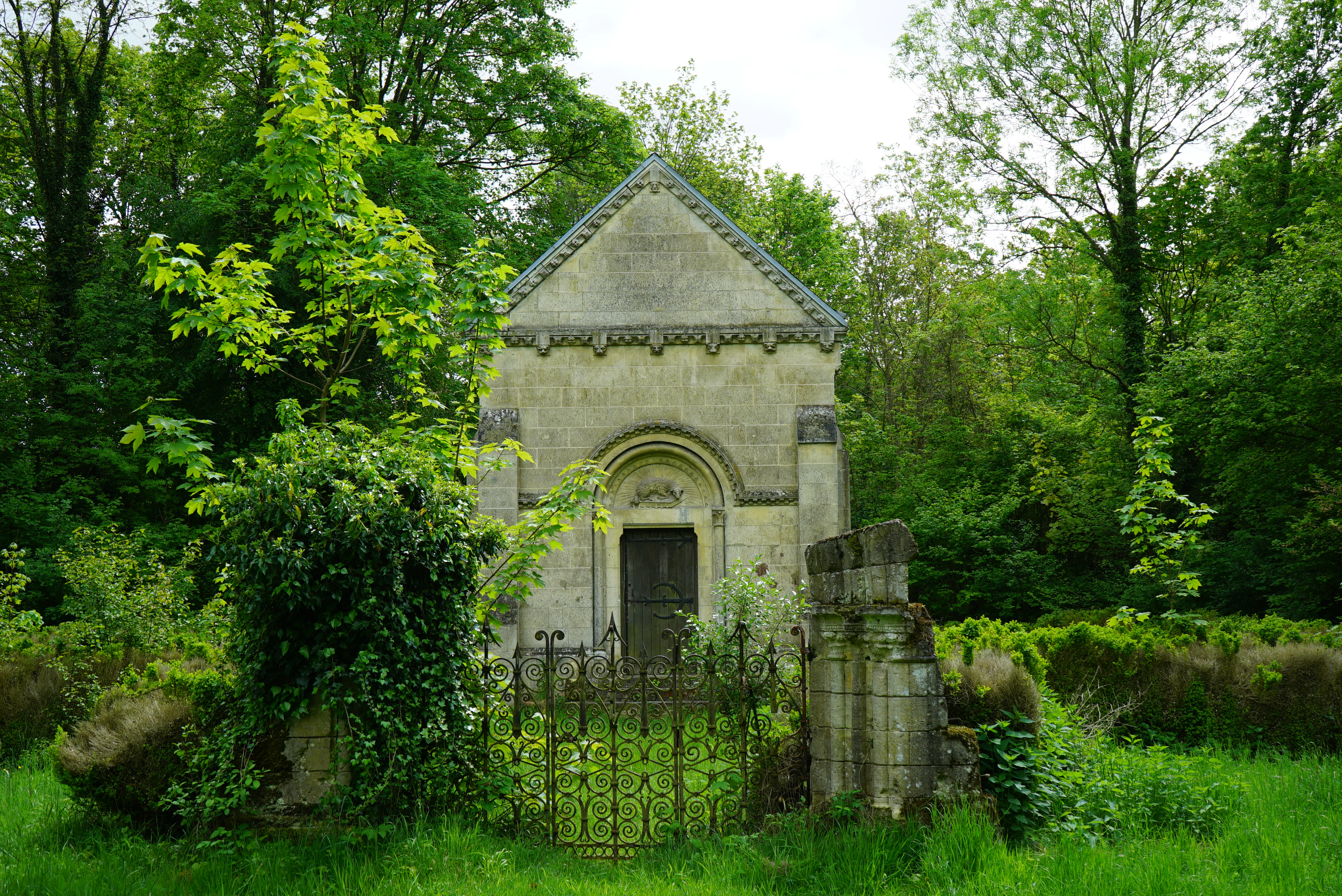 A stone chapel with an arched doorway sits in a clearing surrounded by a dense green forest, fronted by an old wrought-iron gate and fence.