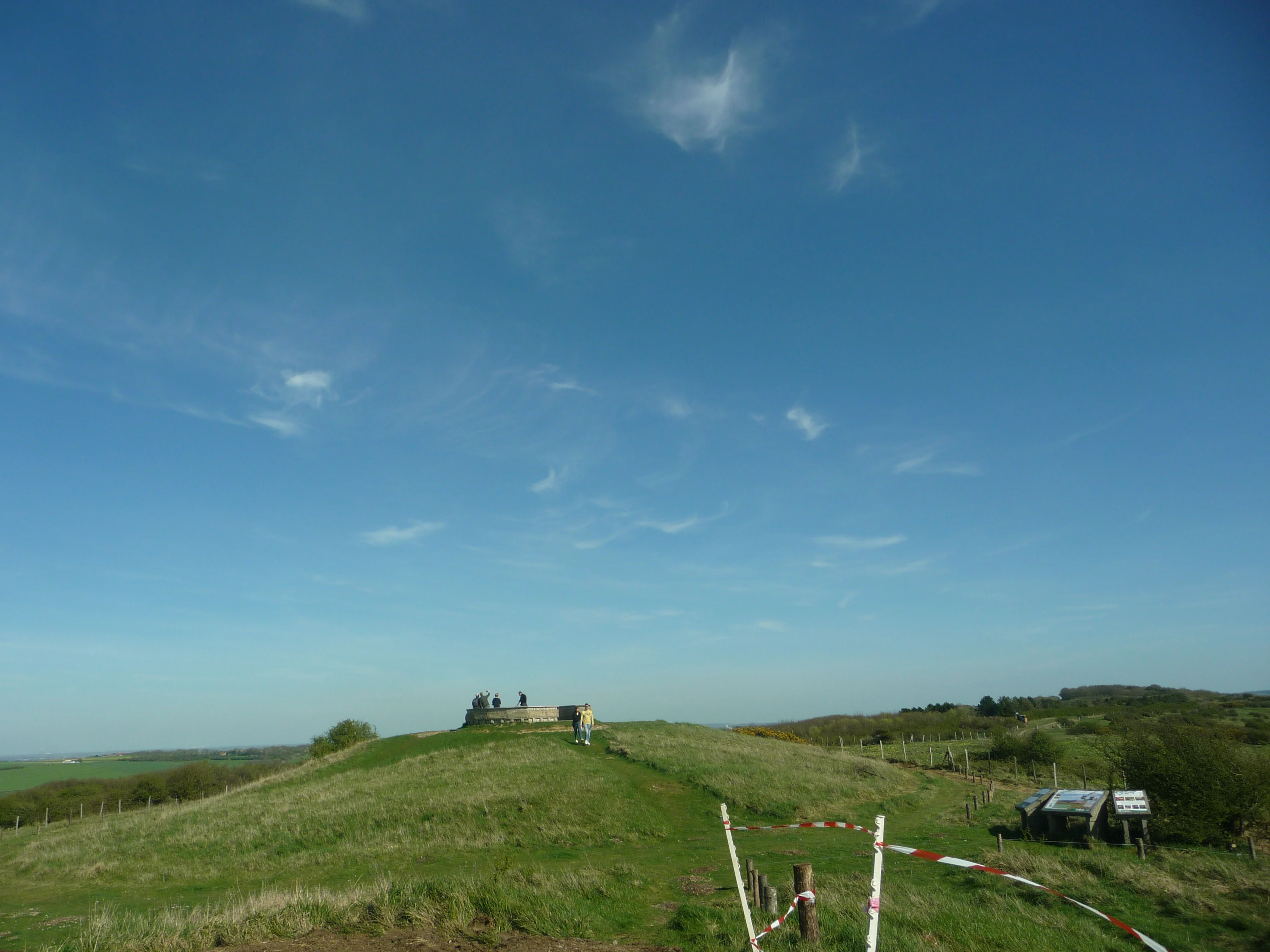 A wide view of a grassy hill summit under a clear blue sky, featuring a stone orientation table structure with small figures of people and a makeshift barrier in the foreground.