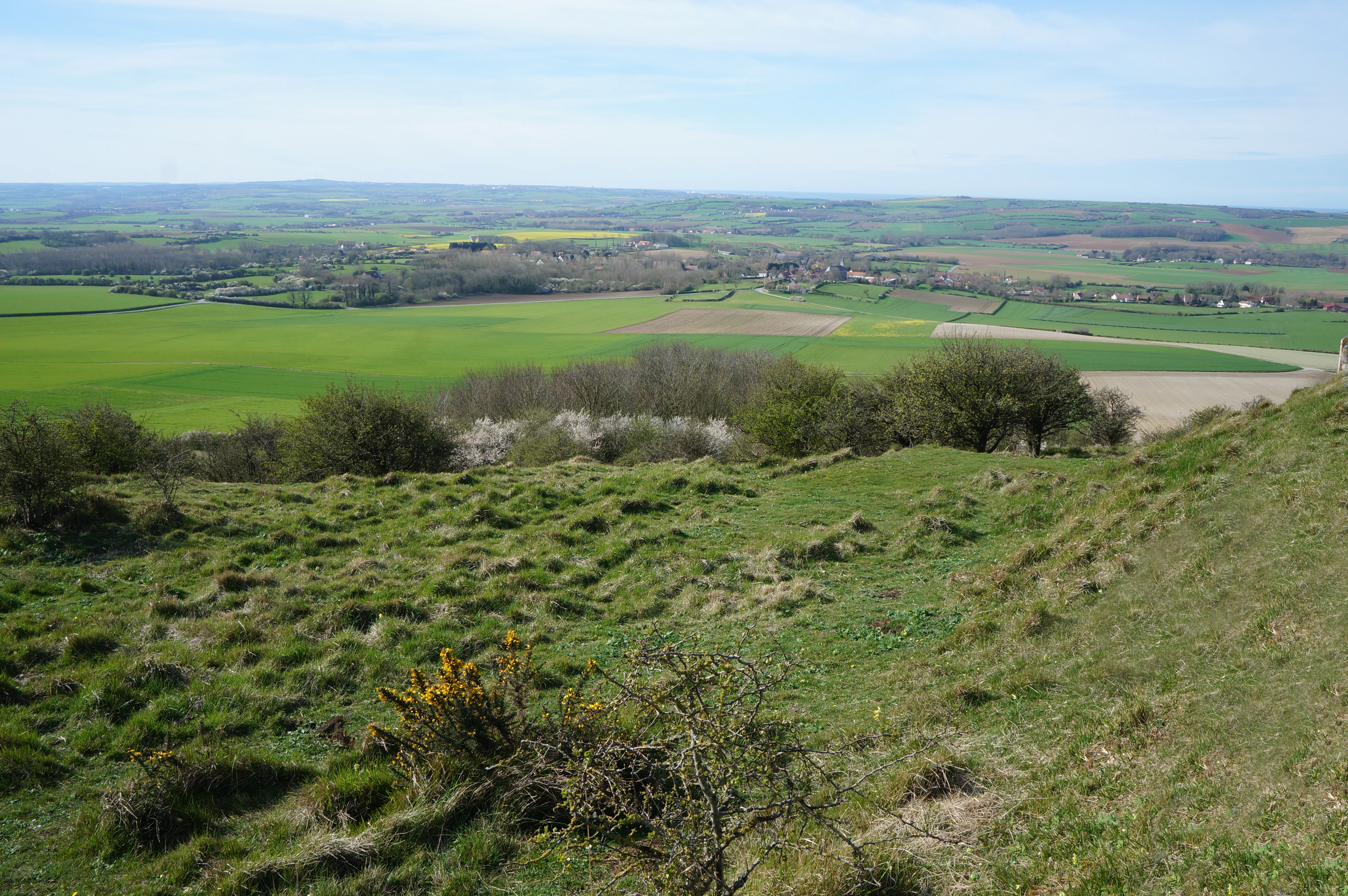 A wide, elevated landscape view from the grassy slopes of Mont de Couple looking out over a patchwork of green agricultural fields, pastures, and small wooded areas under a pale blue sky.