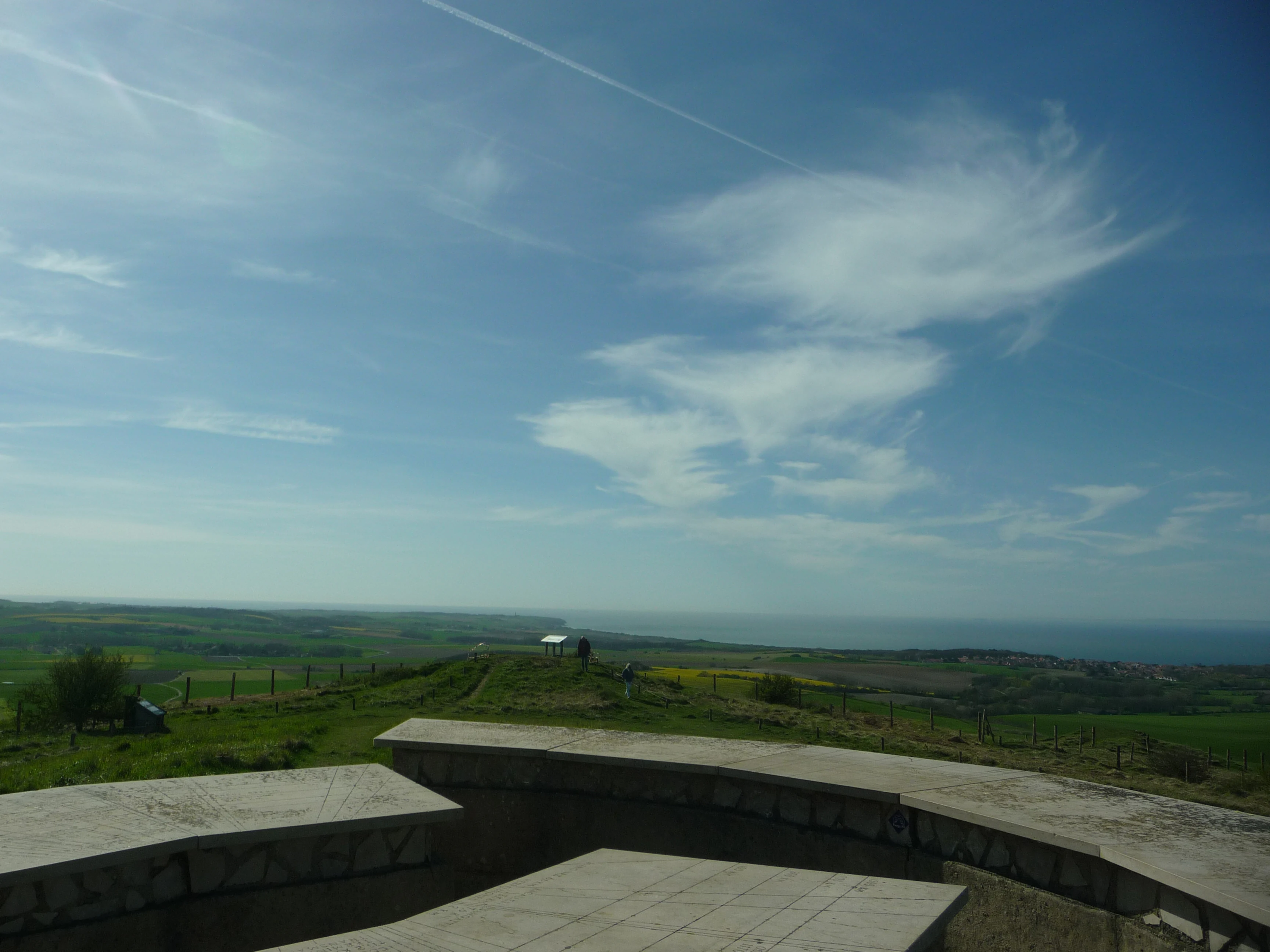 A semicircular stone orientation table sits in the foreground on a grassy summit, looking out toward a distant coastline under a blue sky.