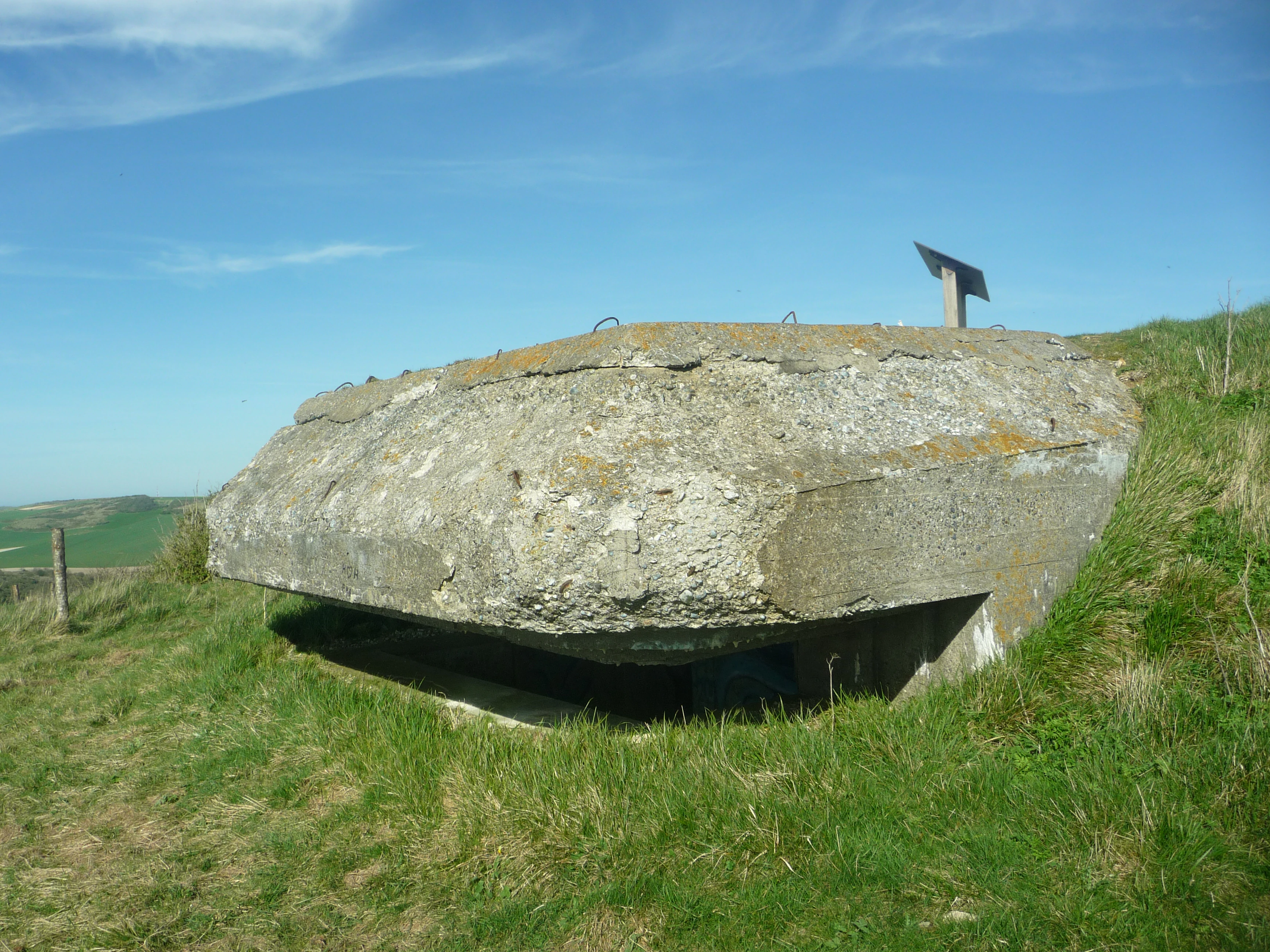 A weathered concrete bunker built into a grassy hillside under a clear blue sky, with a small informational sign visible on top.