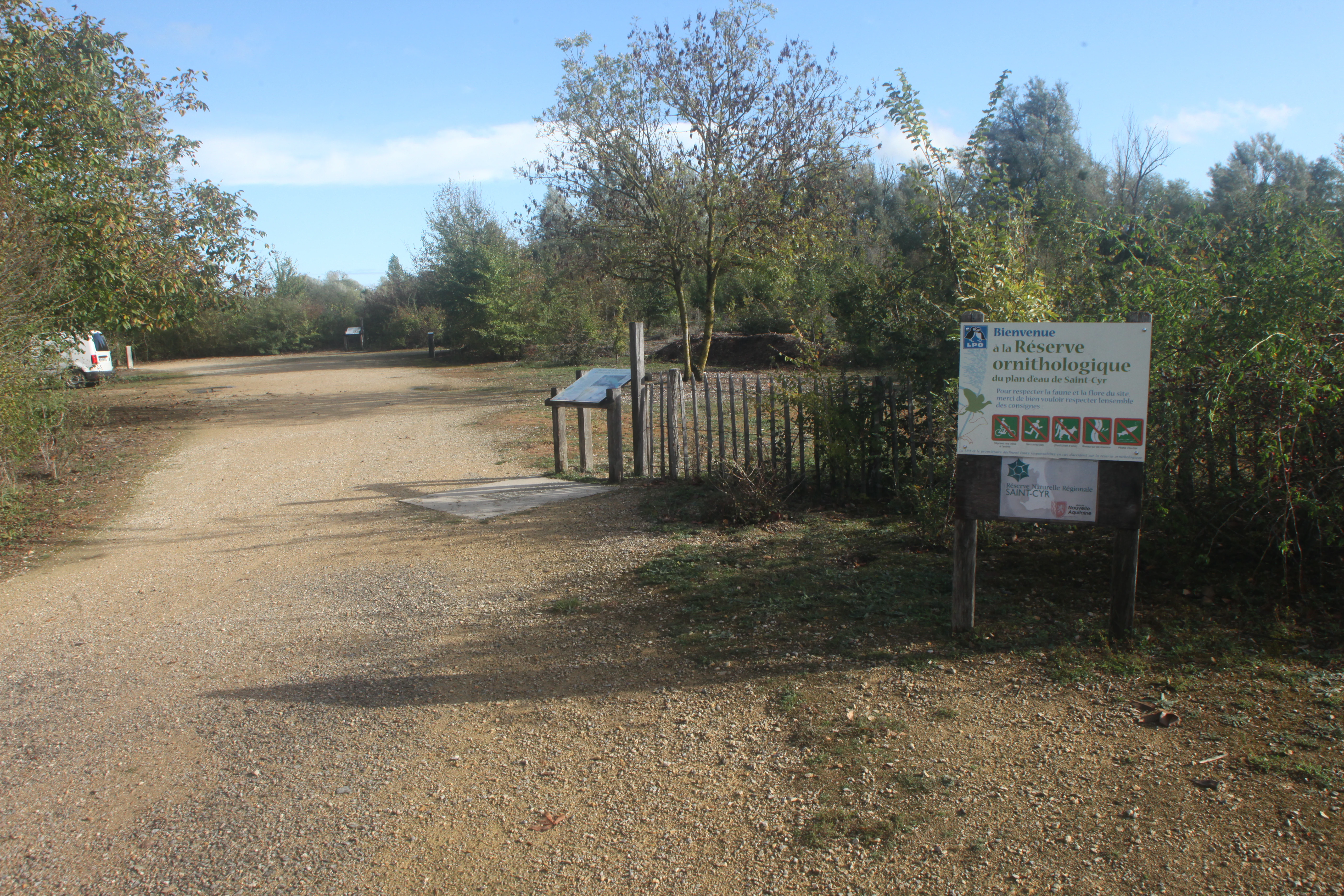 A dirt path leading into a wooded area with an information sign on the right and a wooden fence.