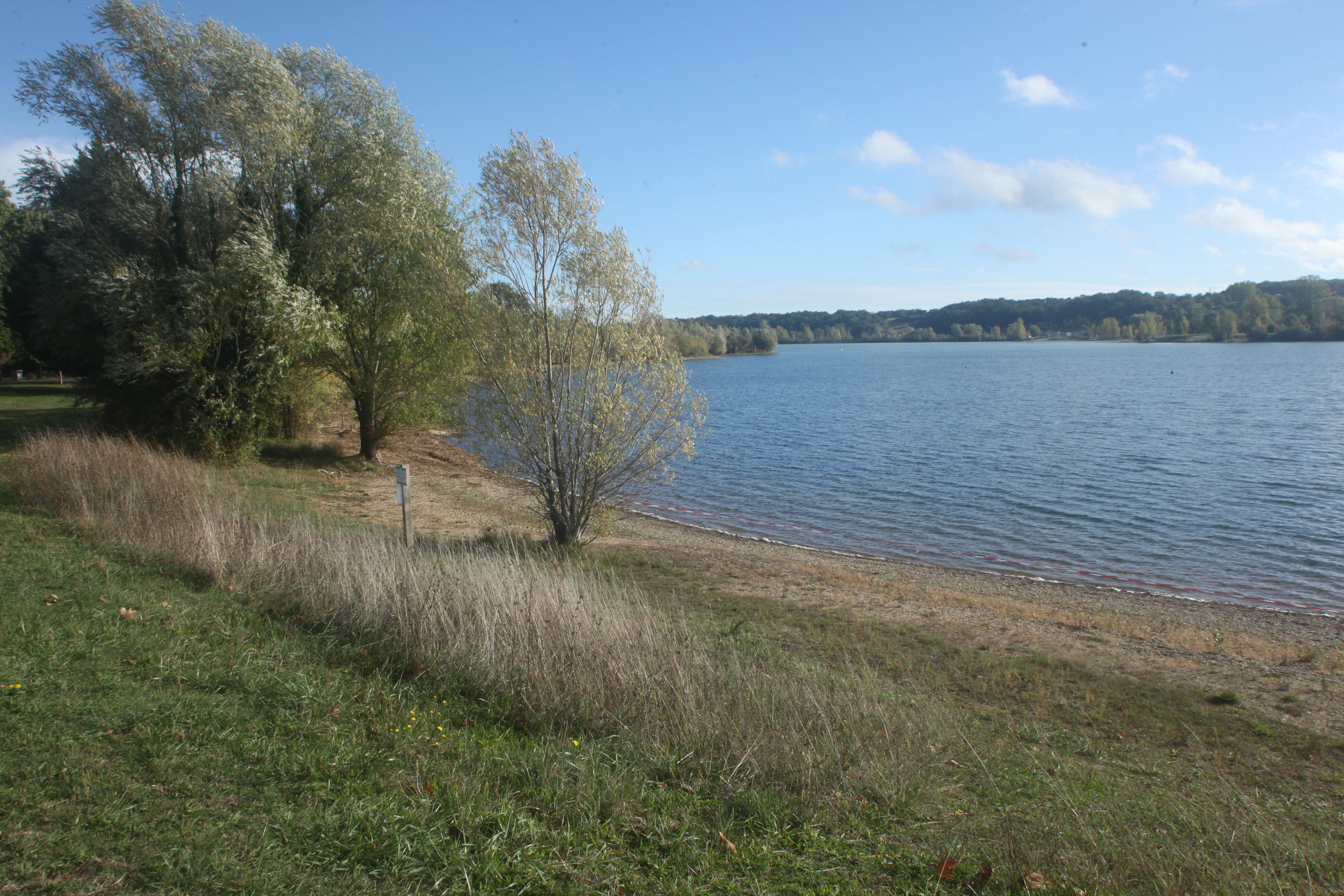 A wide view of a calm lake in the Saint-Cyr leisure park, showing a grassy, sloping bank with trees on the left side and a sandy beach meeting the water.