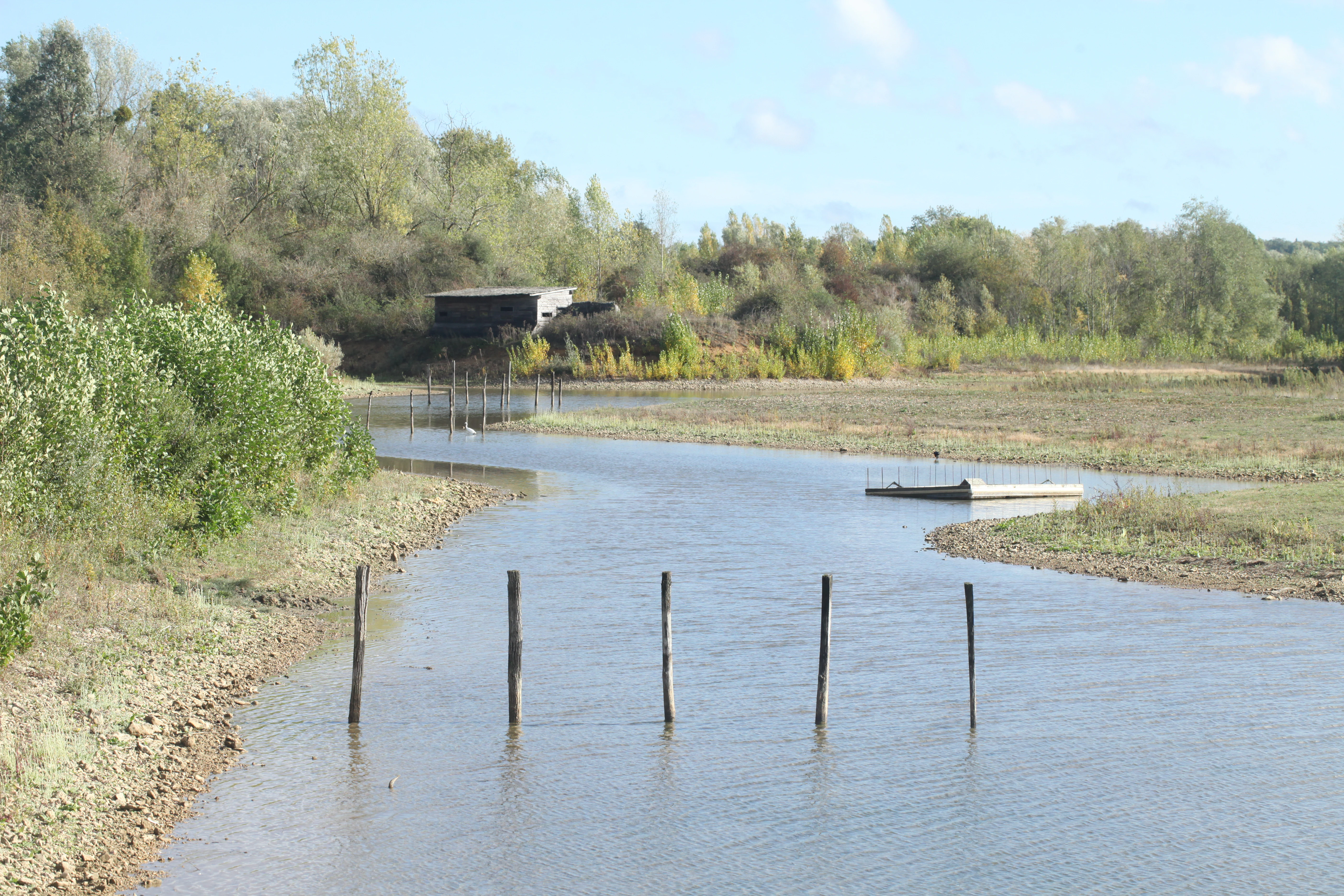 A calm, shallow waterway winding through a nature reserve with a wooden structure on a low hill in the background.