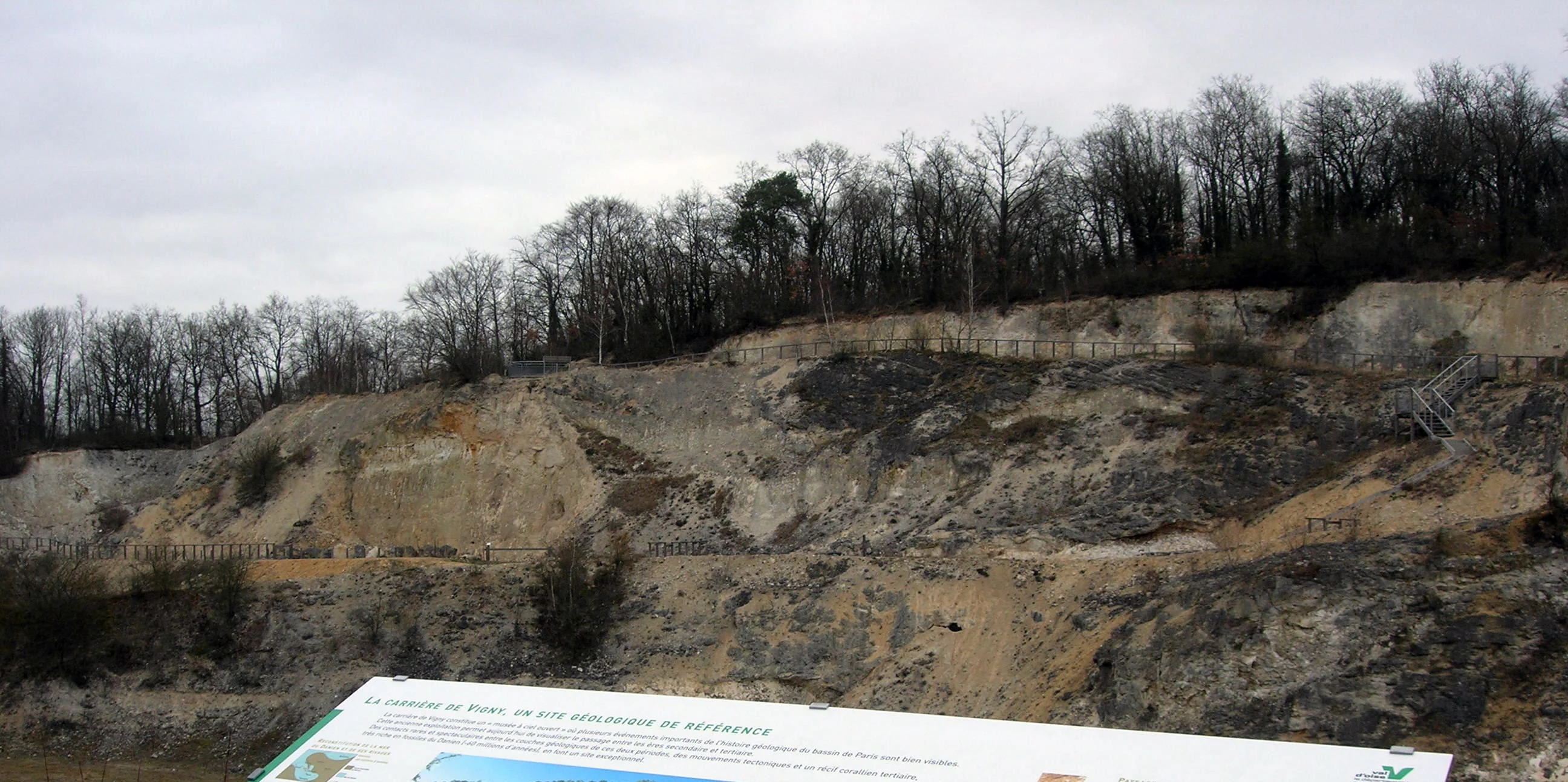 A wide view of a rocky, exposed quarry cliff face under a cloudy sky, with an educational sign in the foreground.