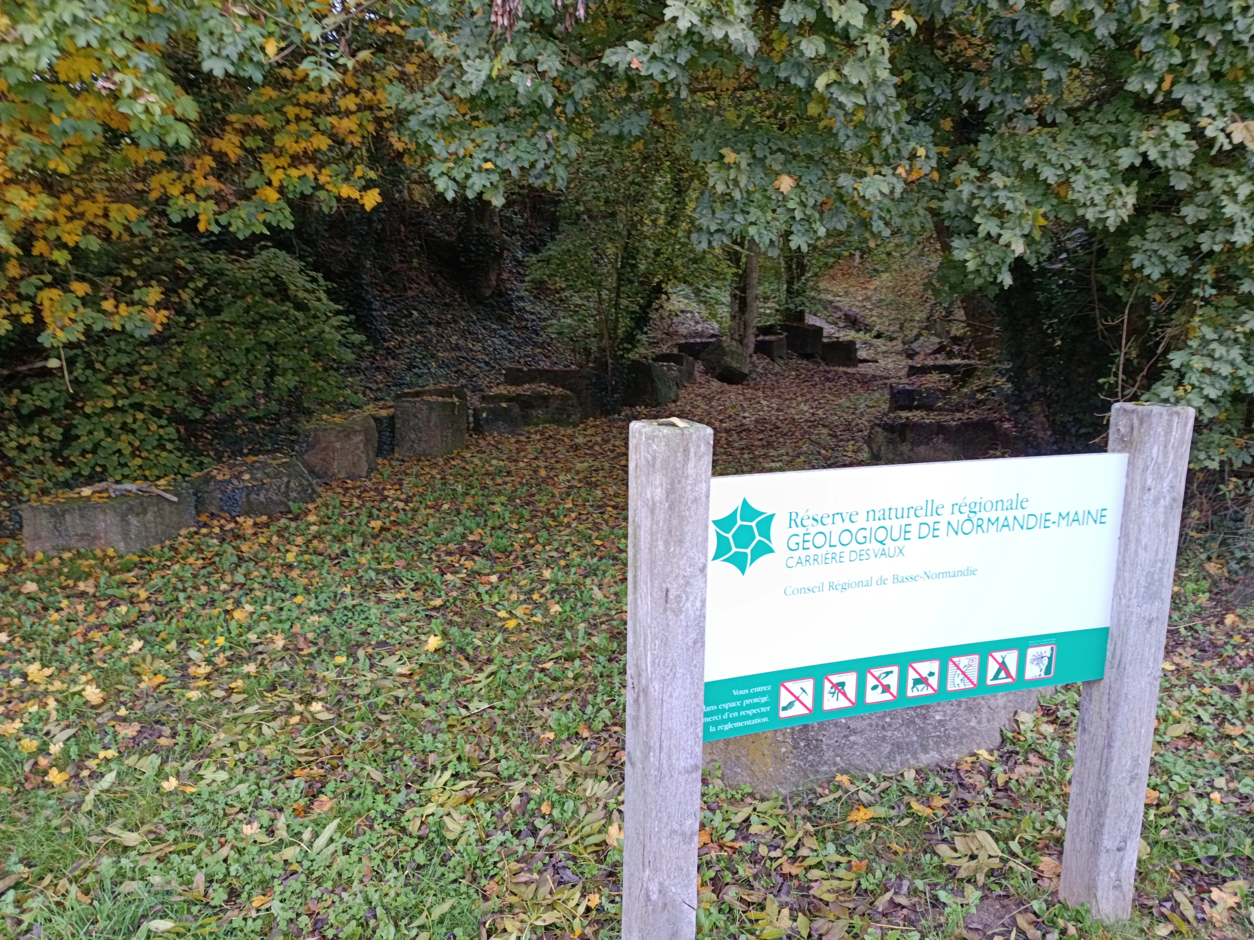 A white and green informational sign reading 'Réserve naturelle régionale géologique de Normandie-Maine Carrière des Vaux' with prohibition symbols, mounted on two wooden posts in a wooded area with autumn leaves on the ground and stone blocks visible in the background.