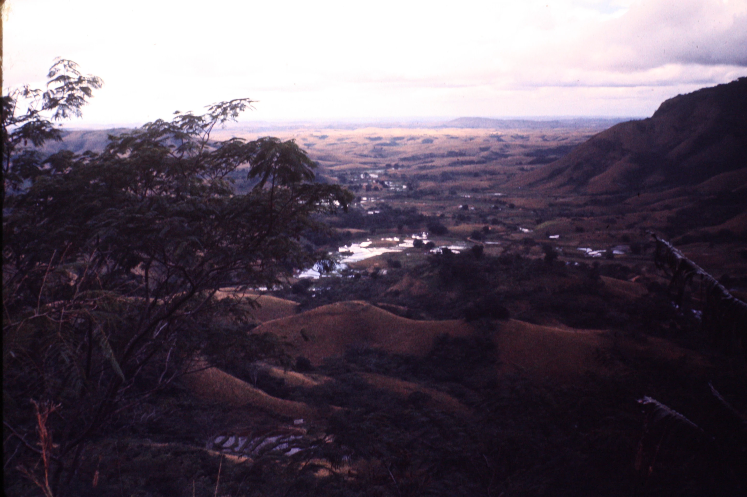A high-angle view looking over a valley with rolling hills, a winding river reflecting light, and distant mountains under a soft light.