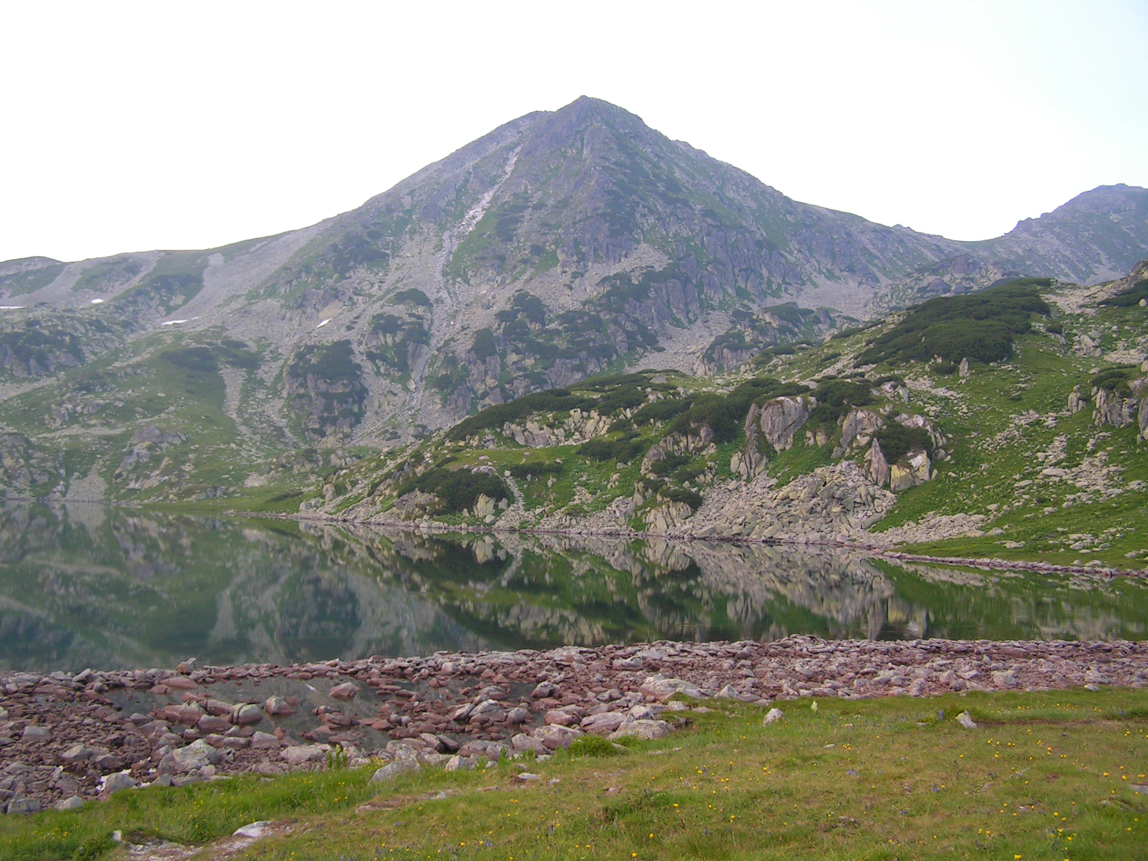 A mountainous landscape with a lake reflecting a rocky peak, surrounded by grassy and rocky terrain