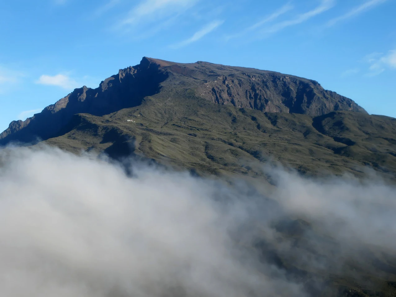 A mountain peak with rocky slopes partially obscured by clouds, under a clear blue sky