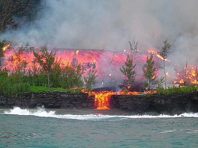 Lava flowing from volcanic eruption into ocean with smoke rising, showing bright orange lava against dark rocks and blue water