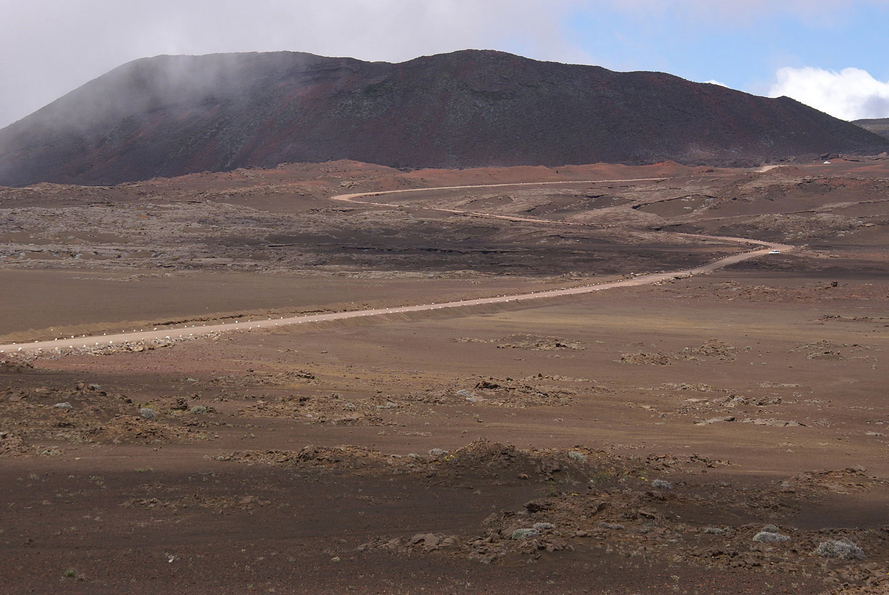 Dirt road traversing arid volcanic plain (Plaine des Sables) with Demi-Piton volcanic cone visible in distant background under partly cloudy sky