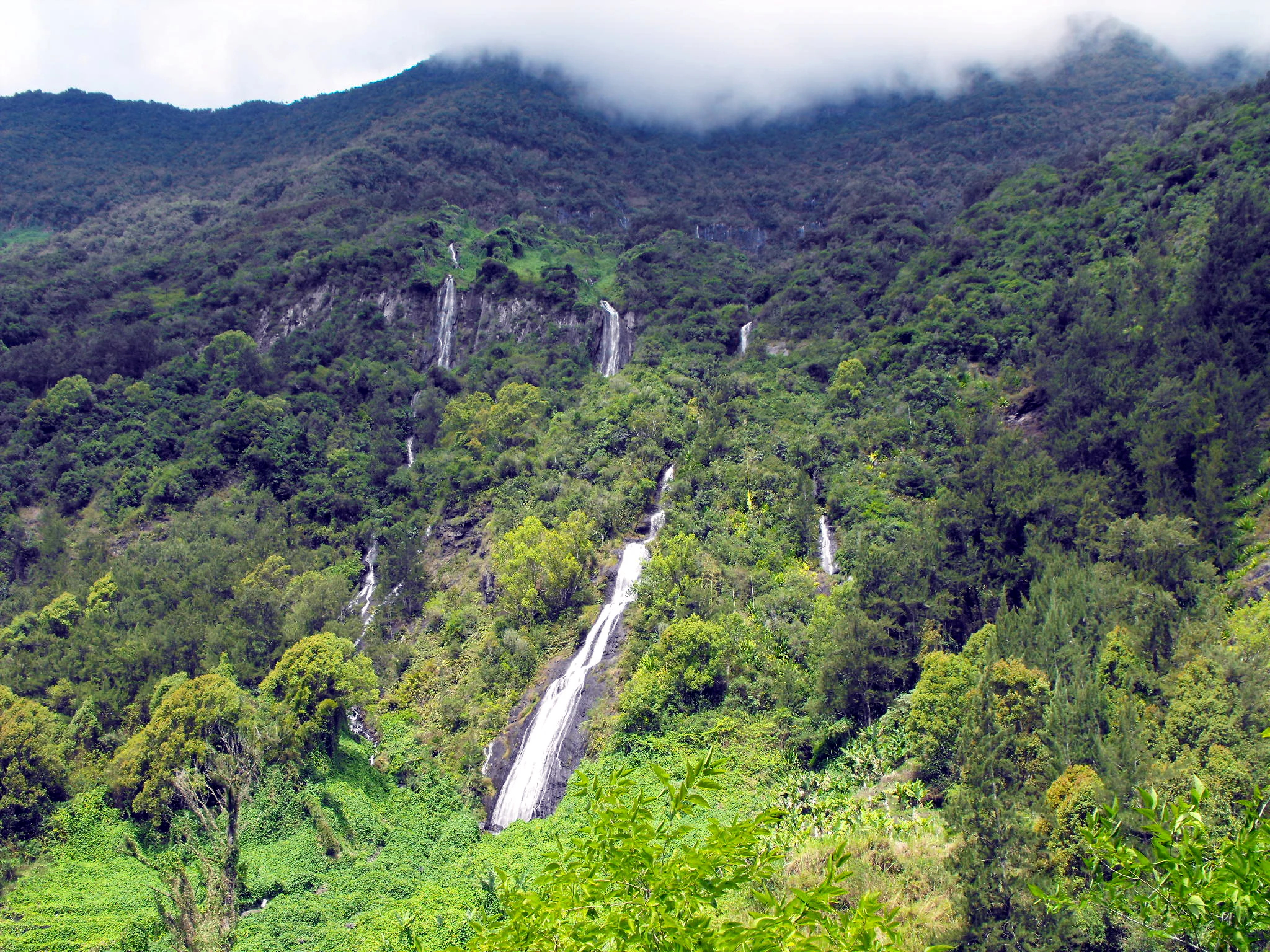 A waterfall cascading down a lush green mountain slope in Réunion National Park, surrounded by dense vegetation with multiple smaller waterfalls visible in the background