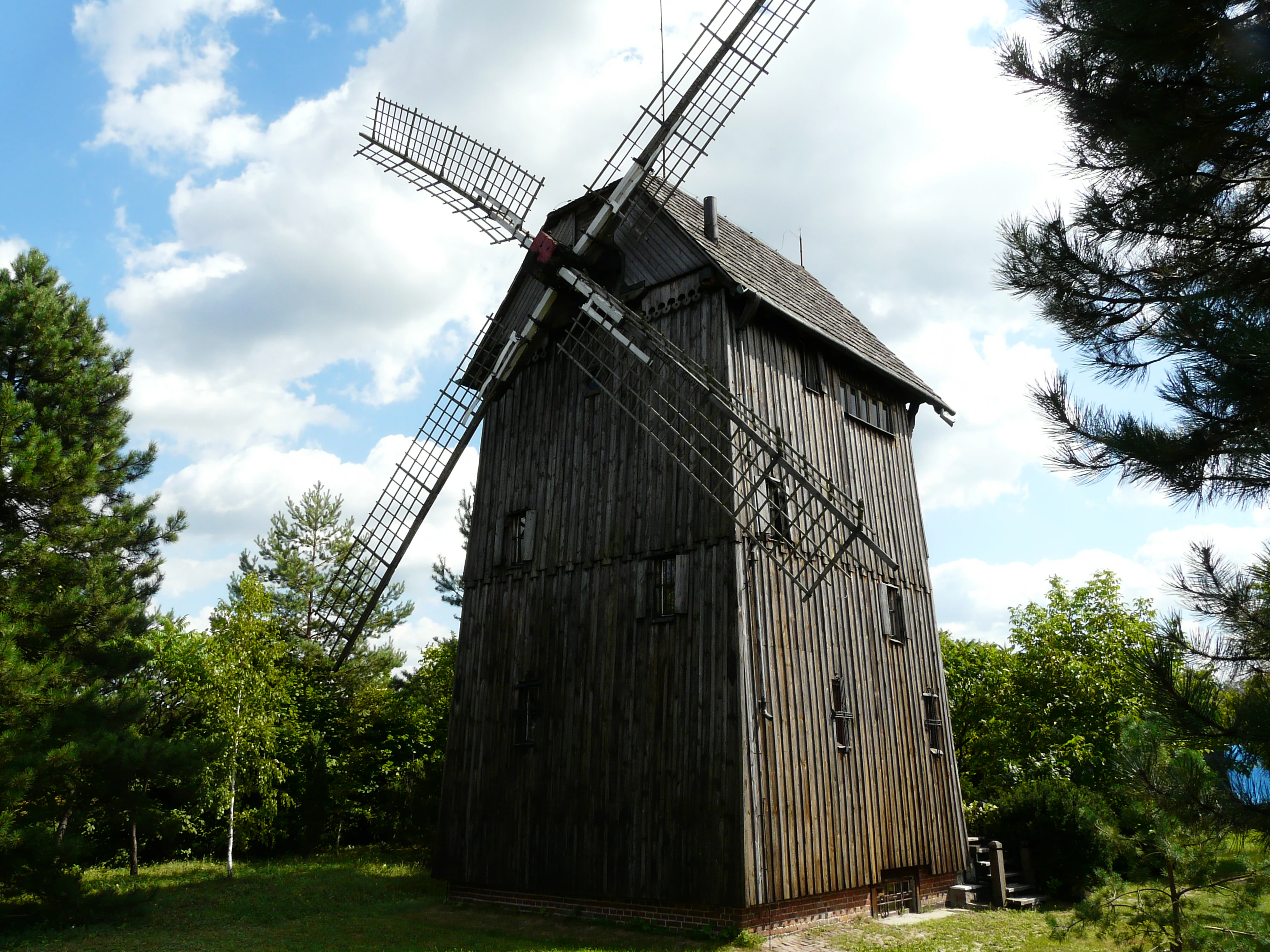 A historic wooden windmill with large blades standing in a green, tree-filled landscape under a partially cloudy blue sky.