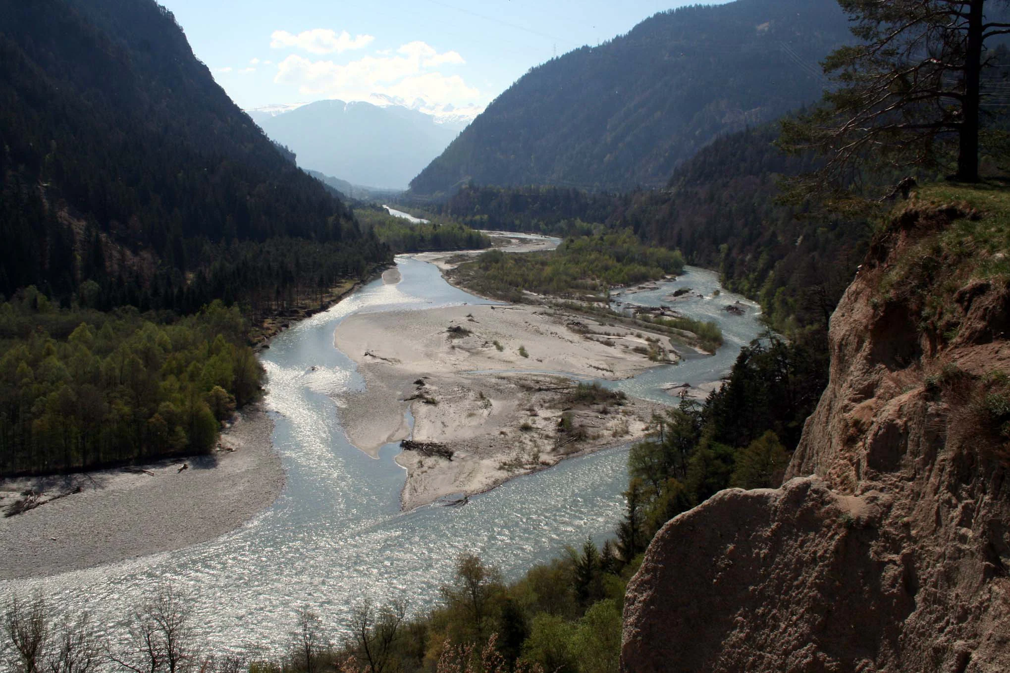 A wide, elevated view of a river with light-colored sandy banks flowing through a forested valley flanked by steep hillsides.
