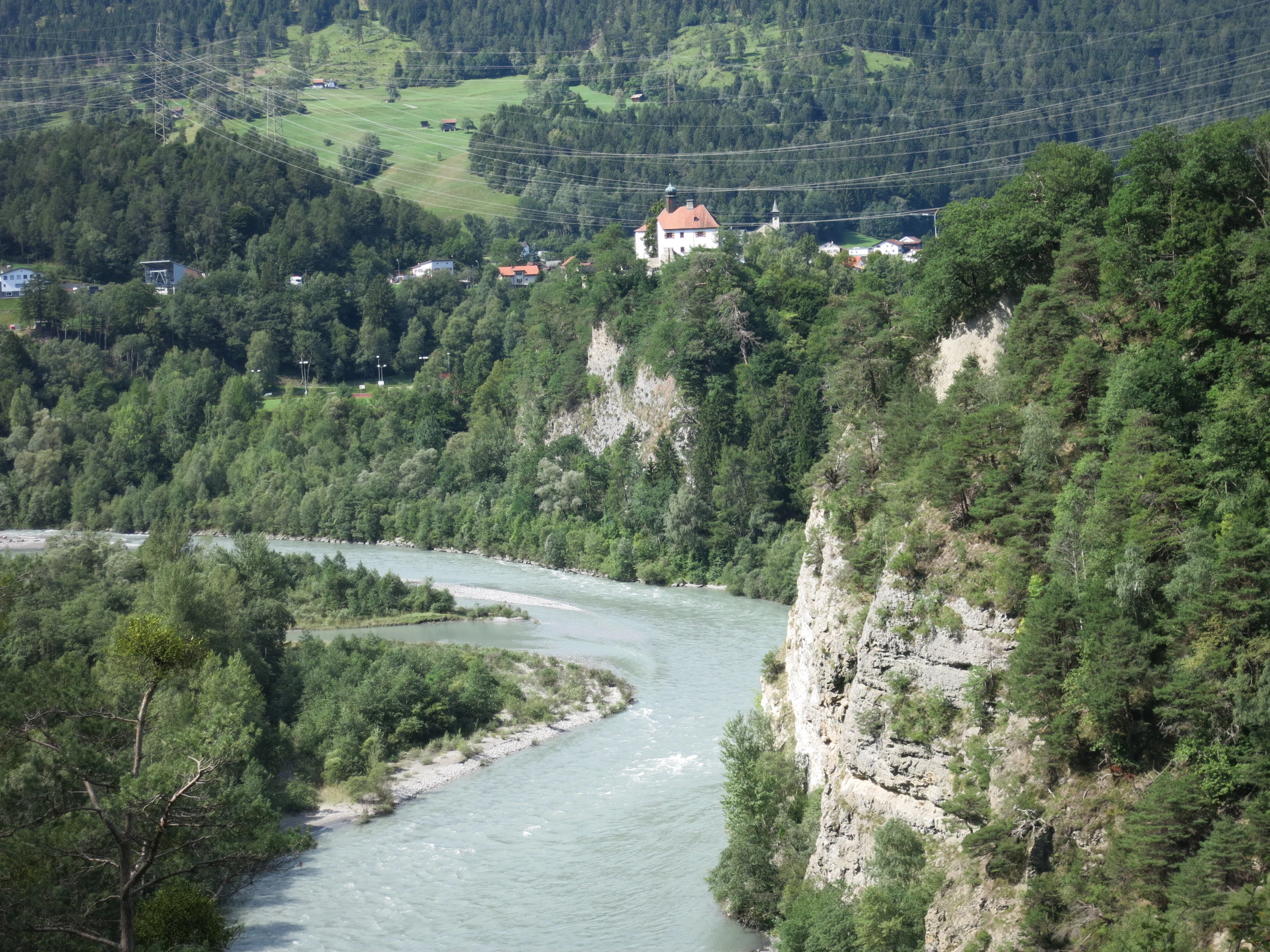 A wide angle view of the Hinterrhein river flowing between forested riverbanks and steep cliffs in the Swiss canton of Grisons, with a building identified as Rhäzüns Castle visible on the hill in the distance.