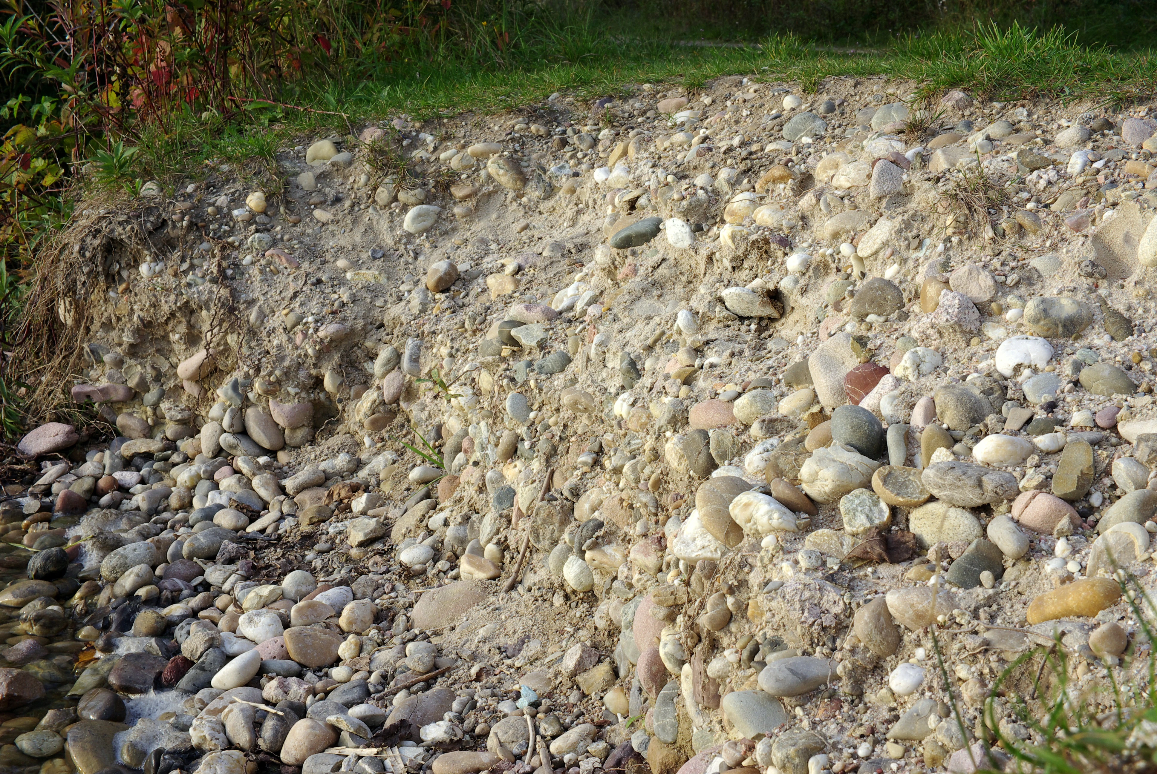 A close-up view of a sloped riverbank composed of mixed gravel, sand, and smooth stones, with a small strip of grass at the top.