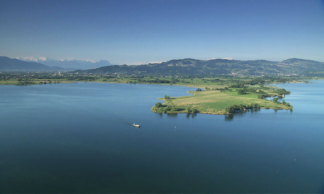 An aerial view of the Rohrspitz peninsula jutting into the calm blue waters of Lake Constance, with forested hills and distant mountains in the background under a clear sky.