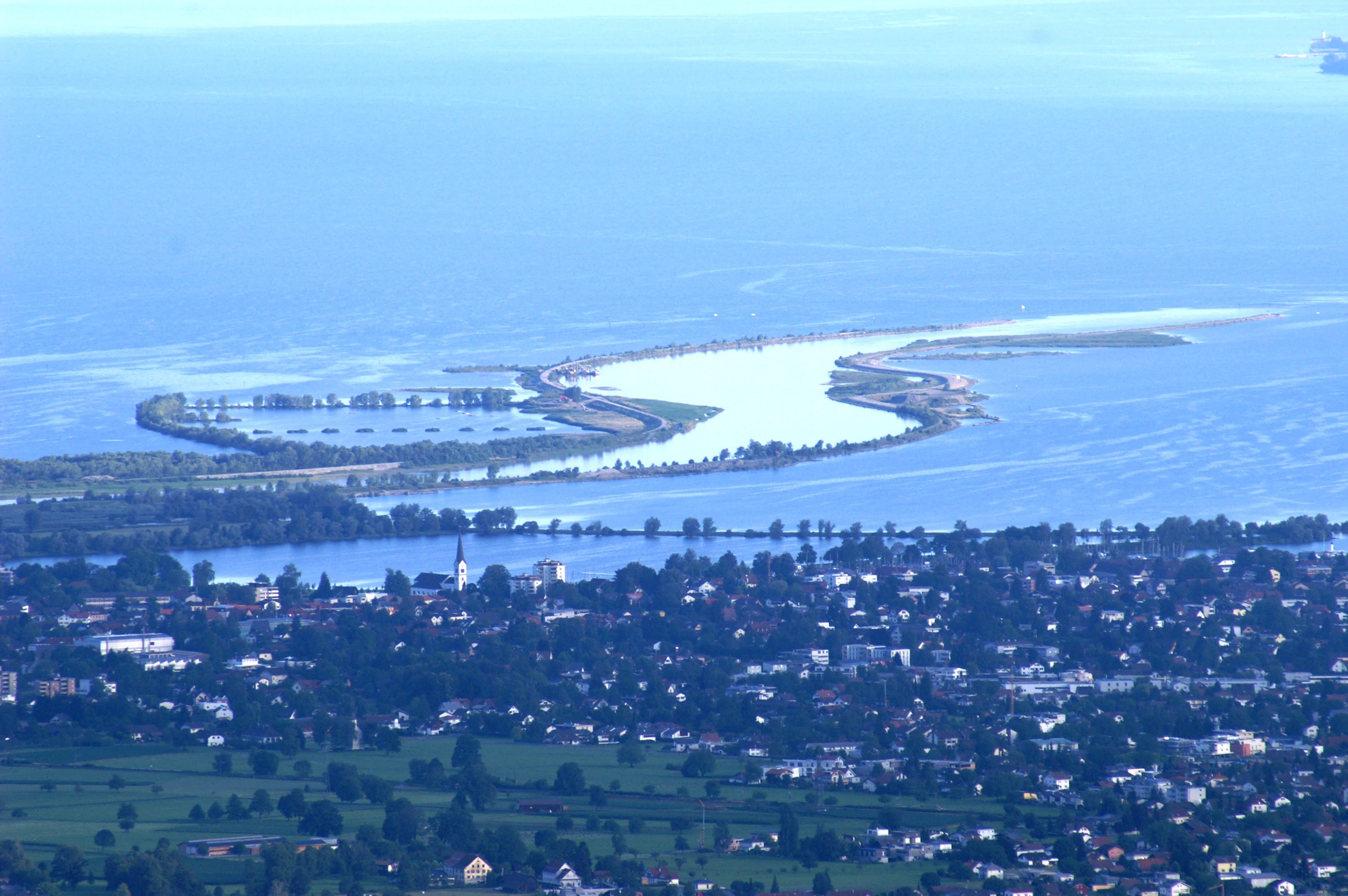 High angle view of the Rhine River flowing into Lake Constance, with the delta shoreline visible and a town in the foreground.