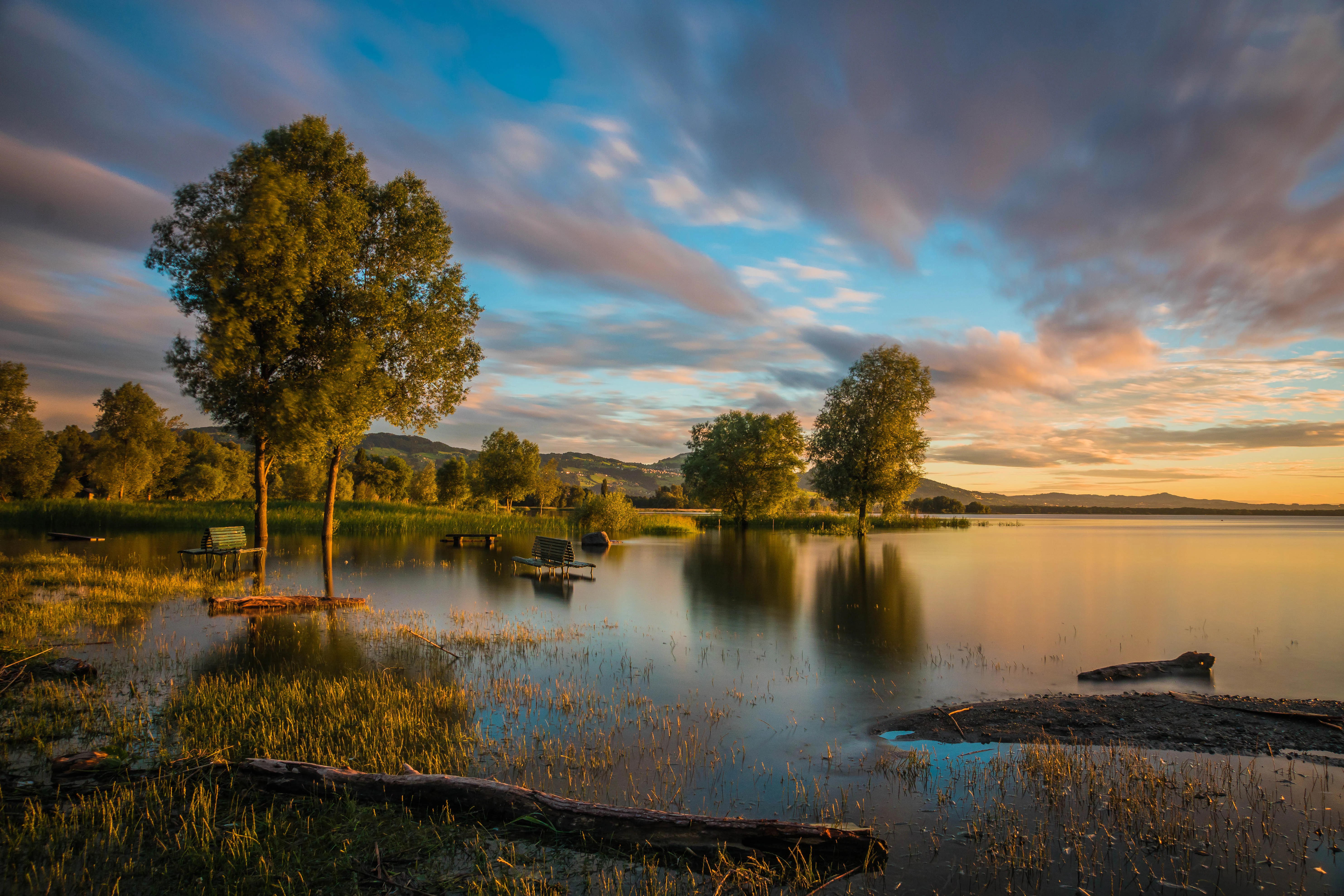 A peaceful lake landscape at sunset featuring trees standing in shallow water, calm reflections, and a colorful, cloudy sky.