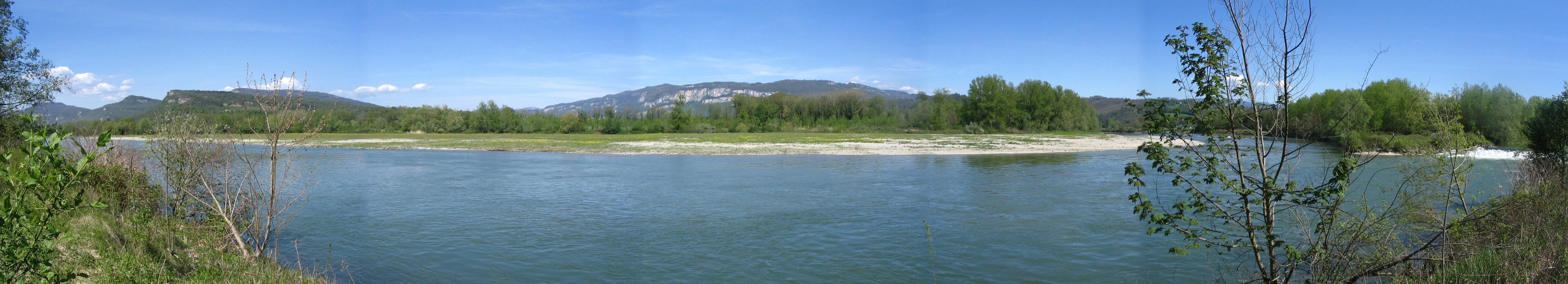 A wide panoramic view of a river confluence with clear blue water, a gravel bank in the center, and green trees along the banks under a clear blue sky.