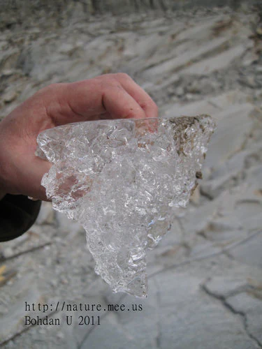 A close up view of a person's hand holding a jagged, translucent fragment of glacial ice against a background of rocky, grey glacial terrain.