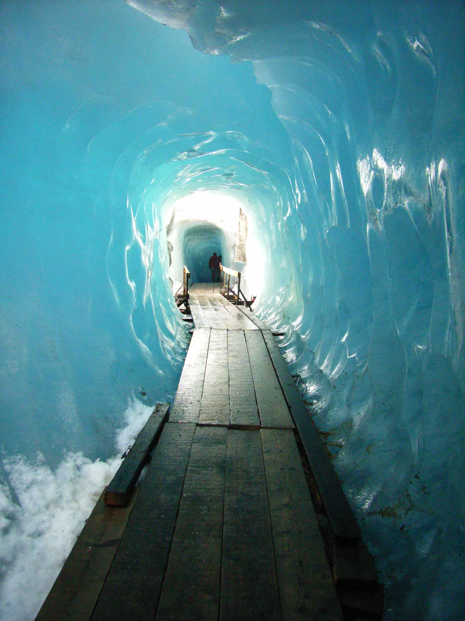 A perspective view inside an ice cave, showing a wooden boardwalk path running through the center of a tunnel surrounded by smooth, translucent blue glacial ice.