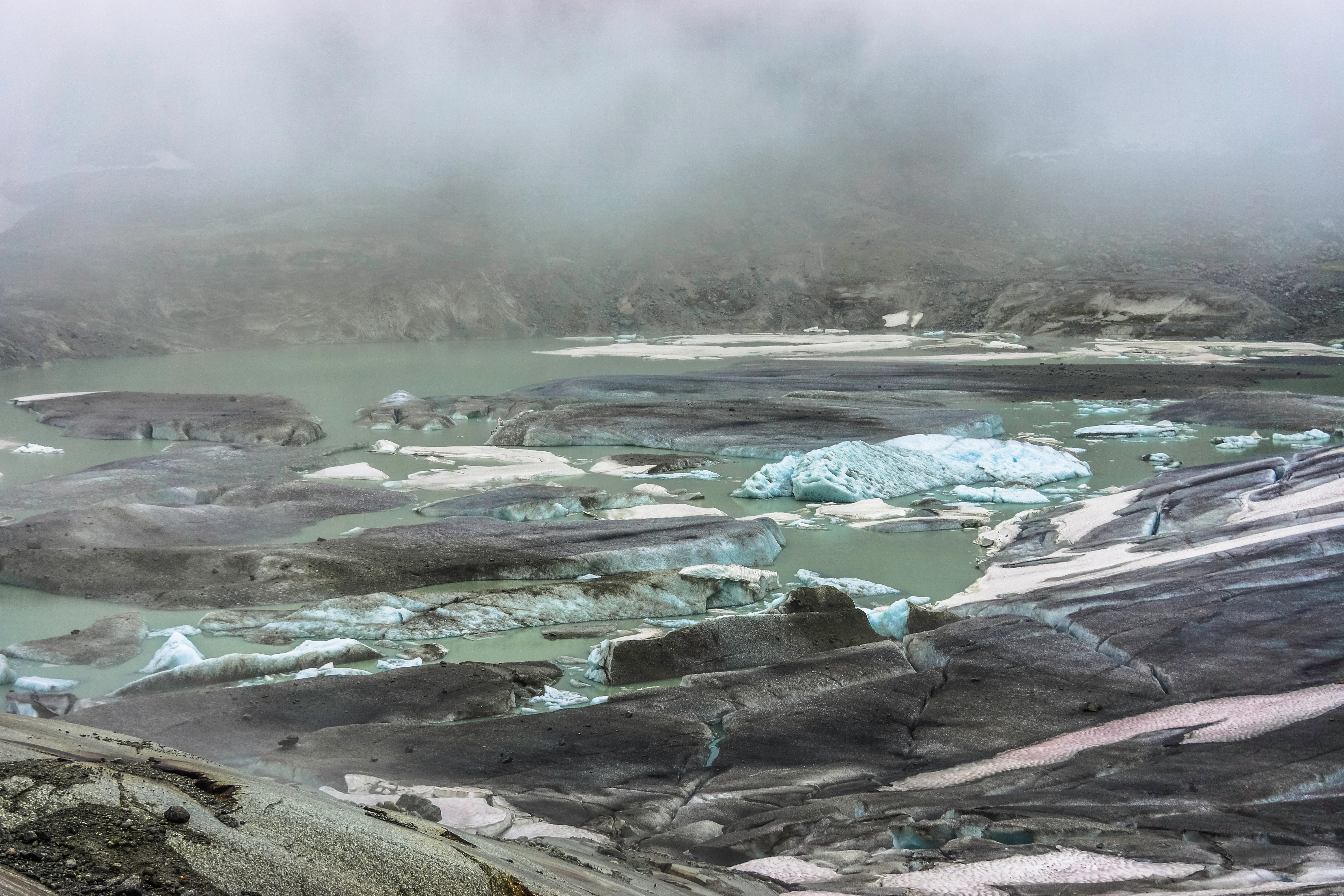 A wide view of a murky green glacial lake containing floating ice chunks in a rocky, barren glacial landscape under heavy mist.