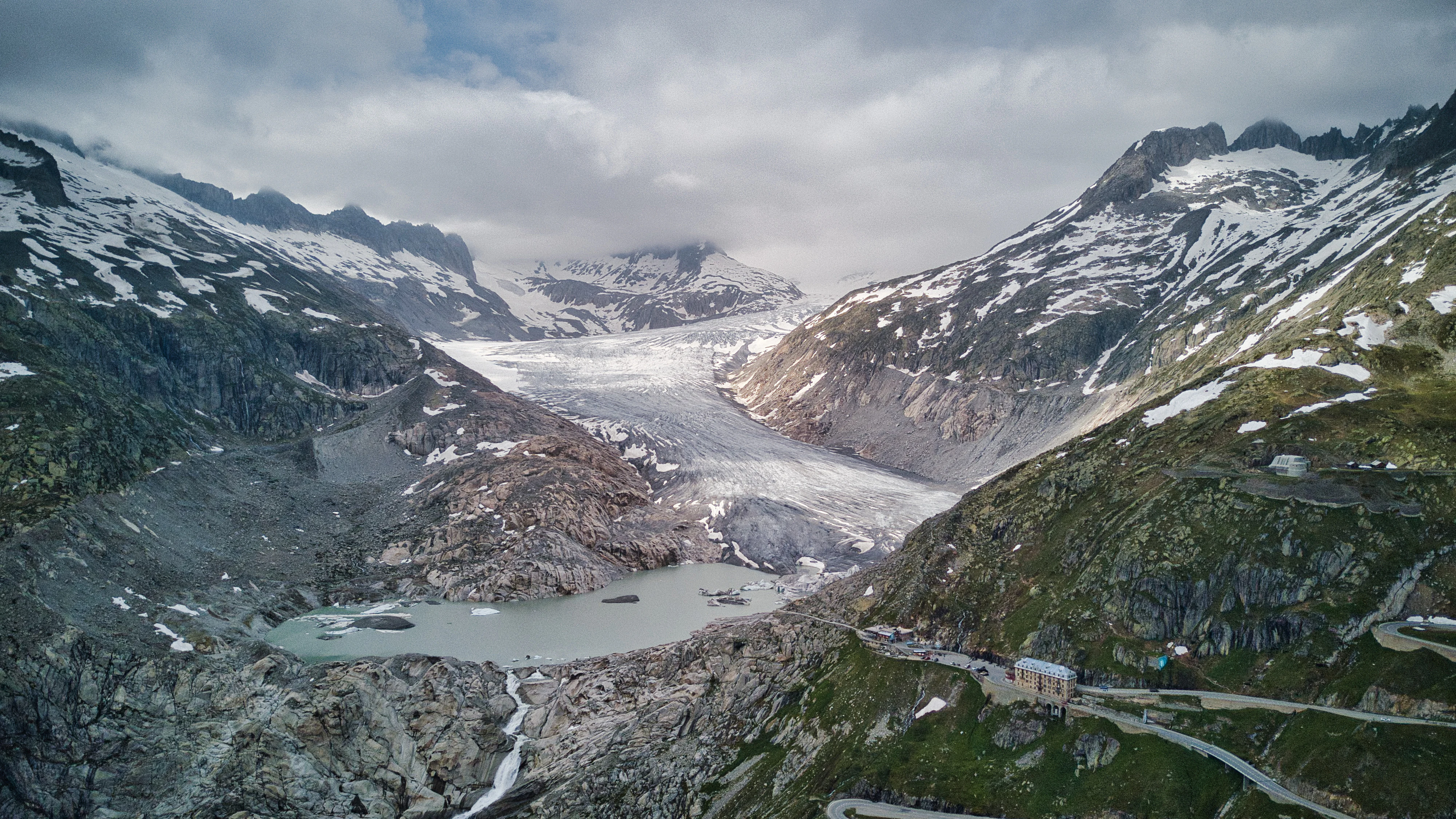 A wide mountain view of the Rhone Glacier flowing down between rocky slopes, with a milky proglacial lake at the base and a winding road visible on the right.