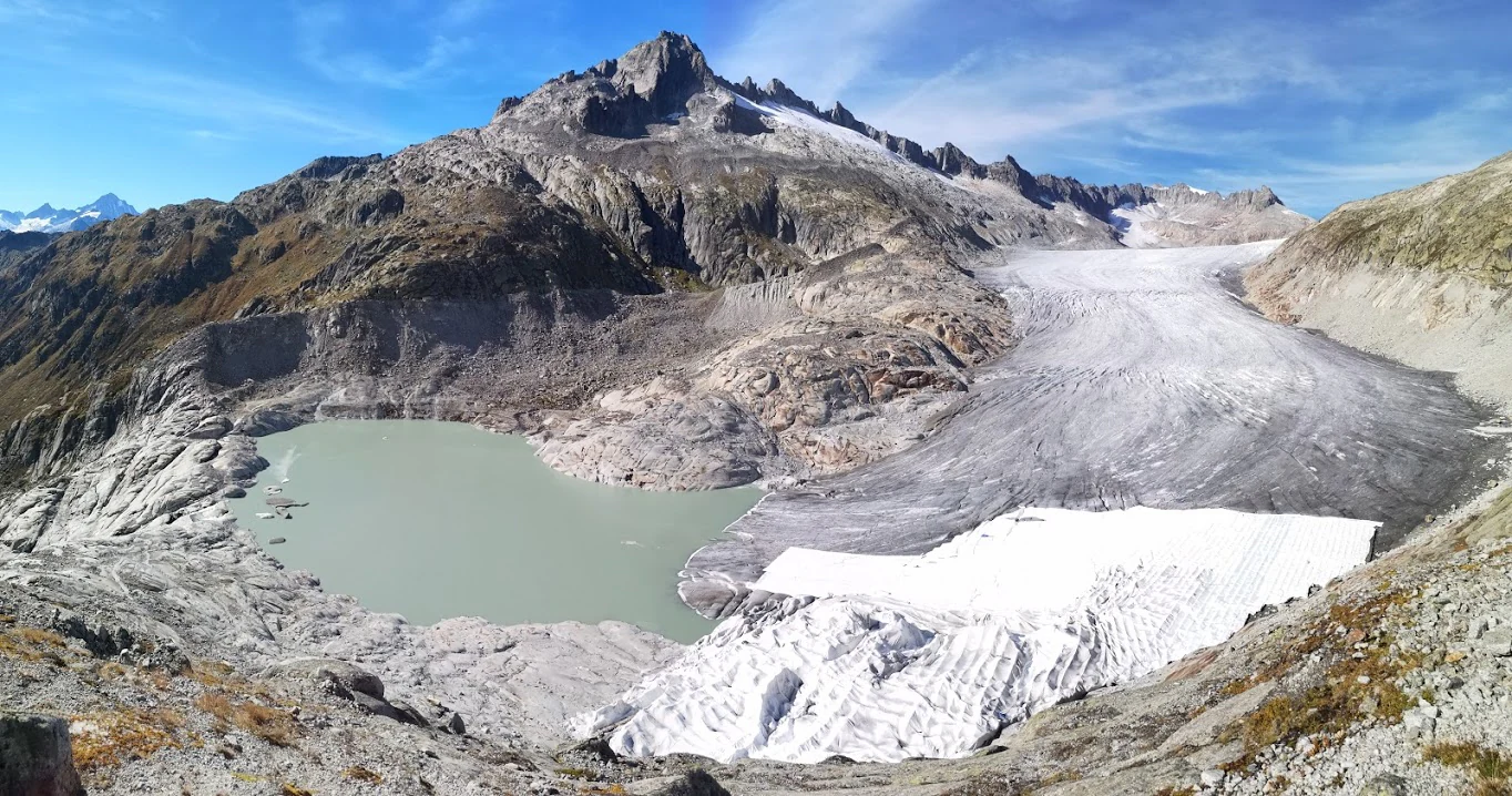 A wide view of the Rhone Glacier featuring a large, light-green glacial lake in the foreground, with the massive ice tongue extending into the center and rocky mountain slopes surrounding the basin.