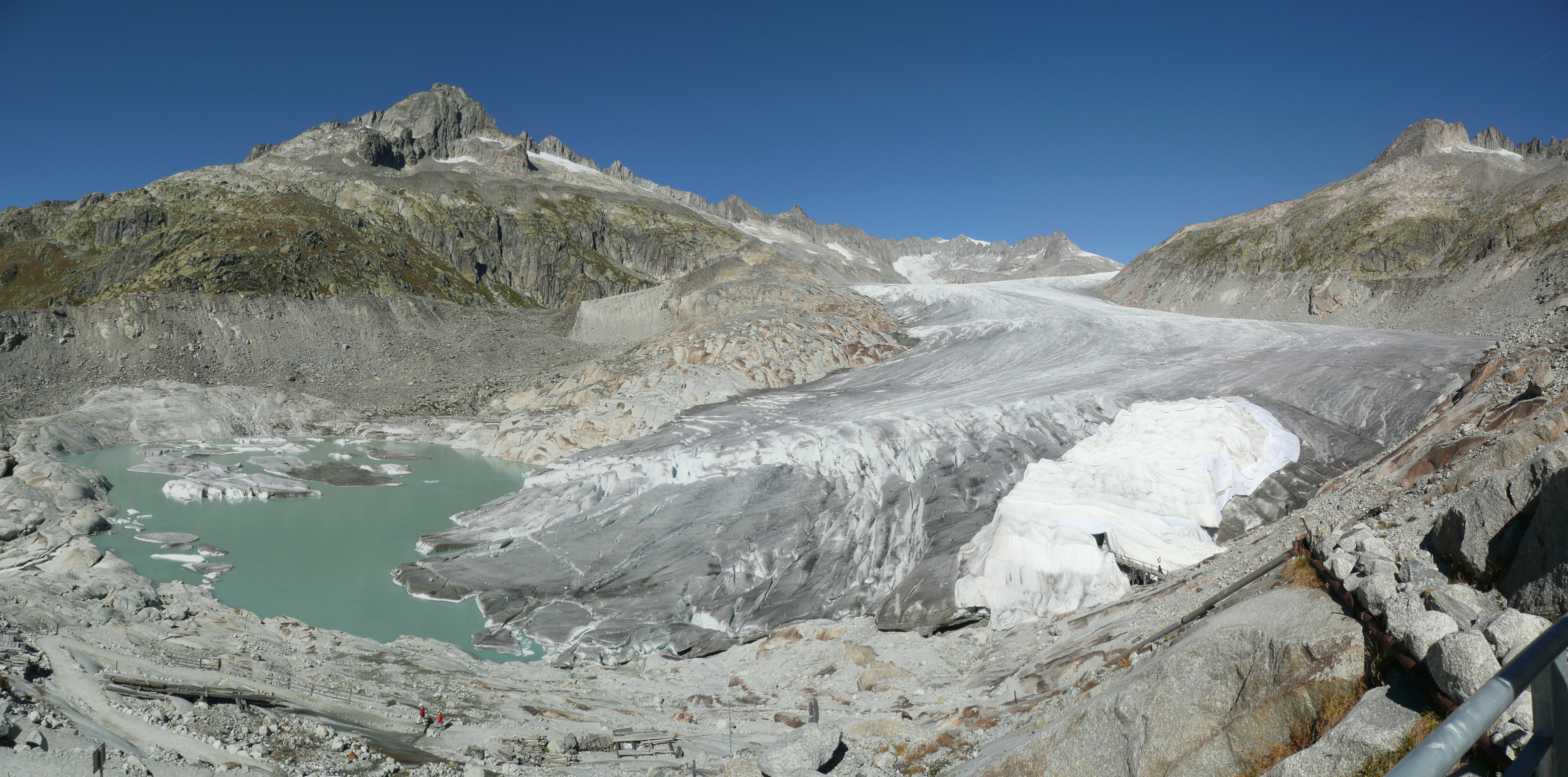 Panoramic landscape of the Rhone Glacier showing a large ice mass receding into a rocky, mountainous terrain with a turquoise proglacial lake in the foreground under a clear blue sky.