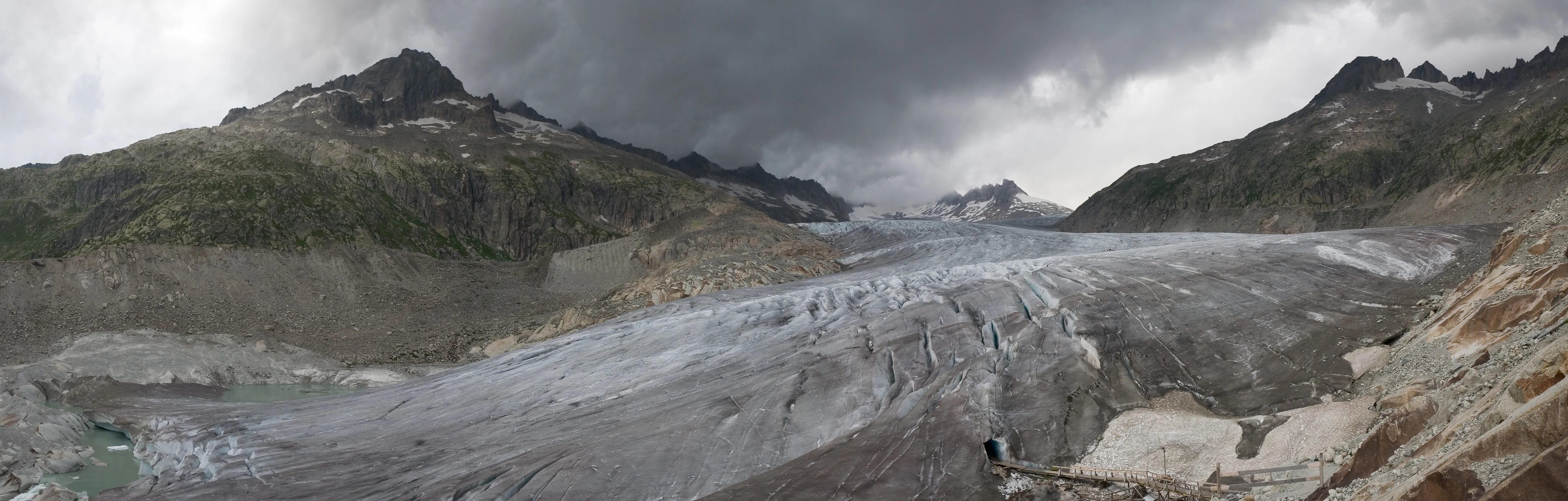 Panoramic photograph of the Rhone Glacier in Switzerland, showing a large, crevasse-filled glacier flowing down a valley between steep, dark, rocky mountain slopes under a moody, overcast sky.