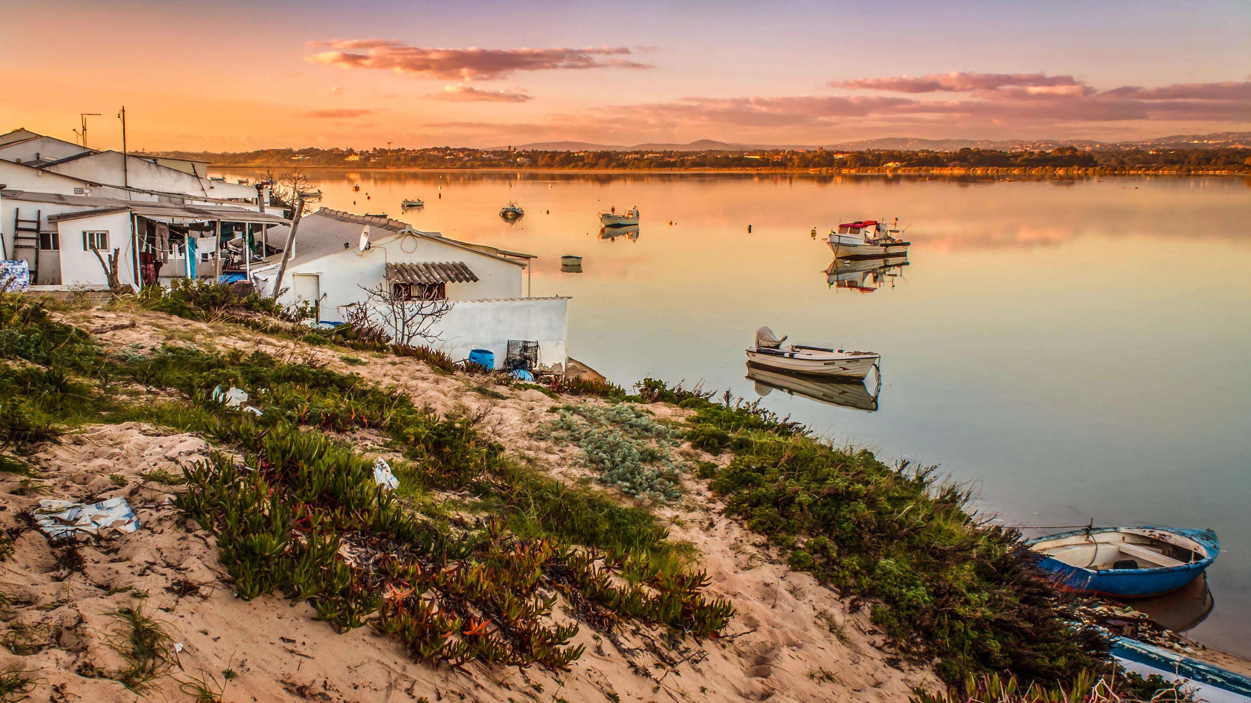 Beachside view of Ilha de Faro at sunset with boats anchored in calm waters, small white buildings, and sparse vegetation on sandy shore