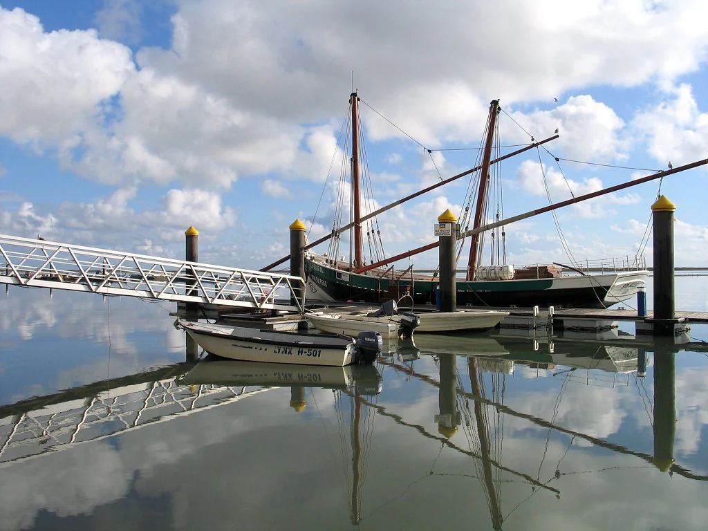 Traditional sailing ship and small motorboats docked at a pier in a calm lagoon with reflections of boats and sky