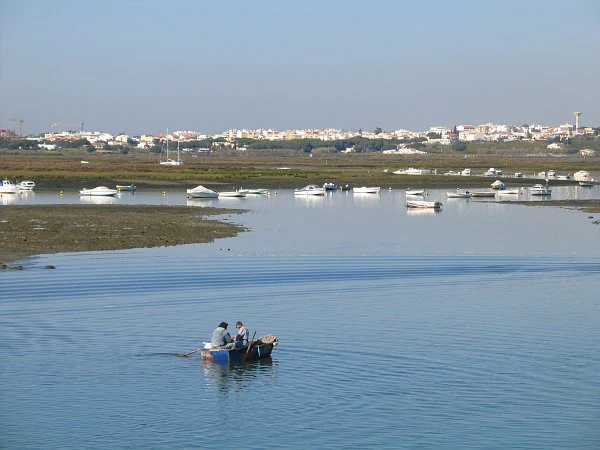 Small boat with two people on calm blue water with boats anchored along the shore and distant buildings on the horizon