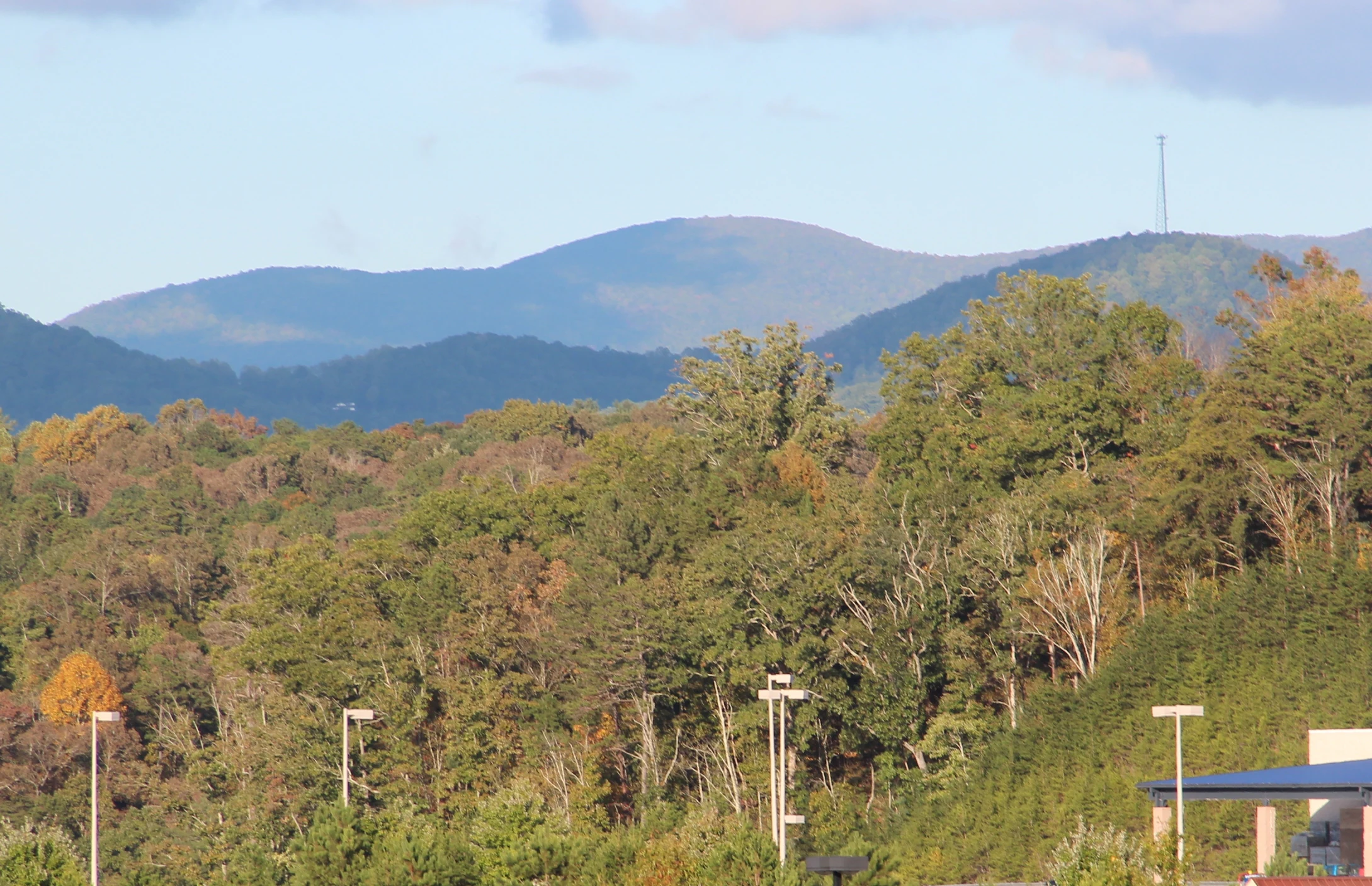 A landscape view of a blue-hued mountain range in the distance behind a dense forest of green and autumn-colored trees, with light poles and a building roof visible in the foreground.