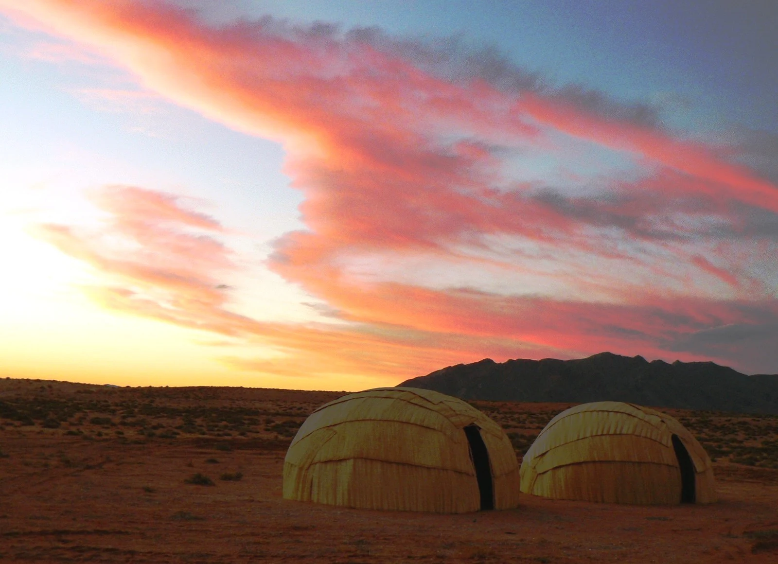 Two dome-shaped, woven traditional dwellings made of reeds sit on flat, arid ground under a vibrant pink and orange sunset sky.