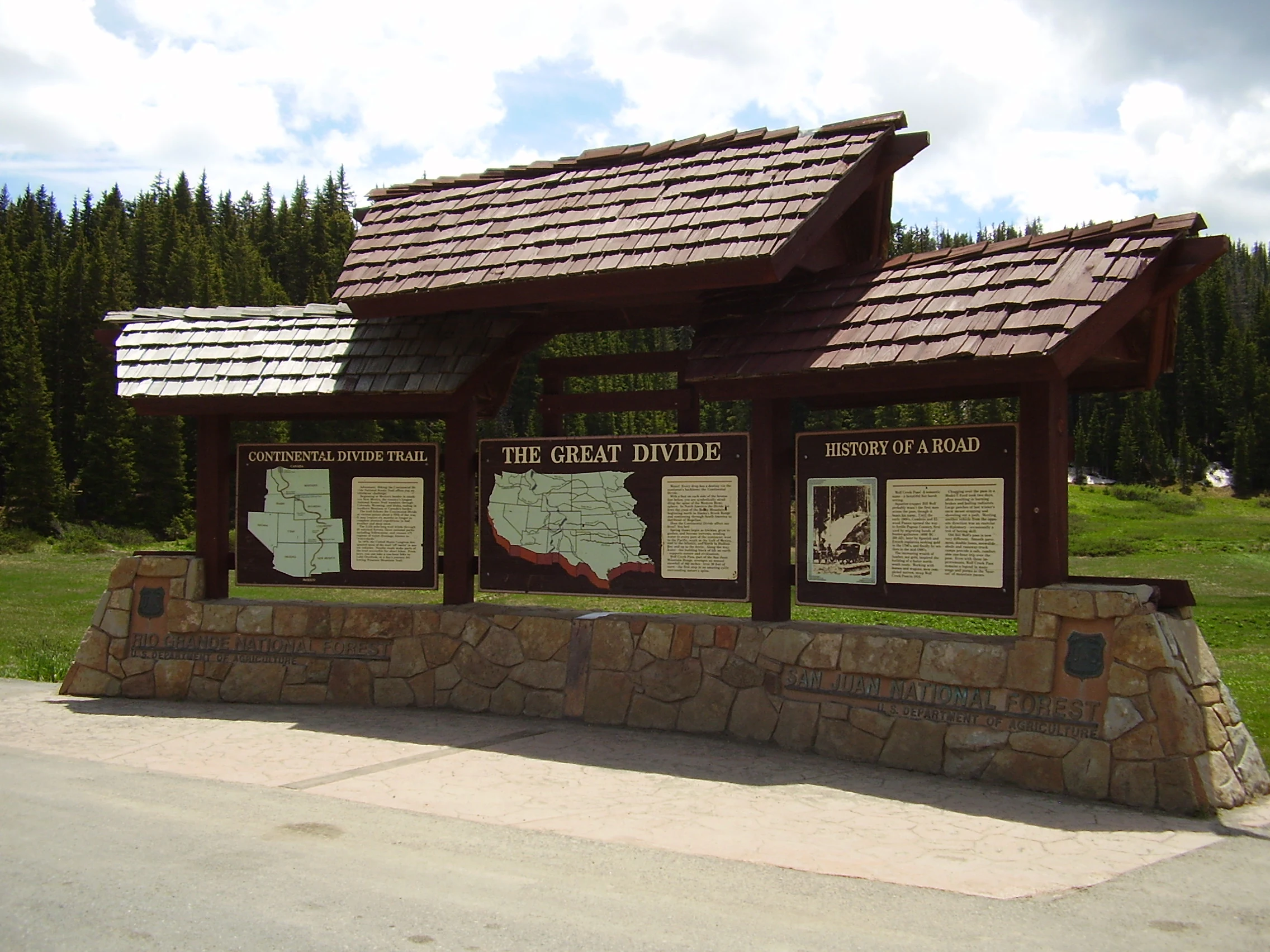 A wooden interpretive sign structure with stone base panels showing maps and text about the Continental Divide, situated in a forested area.