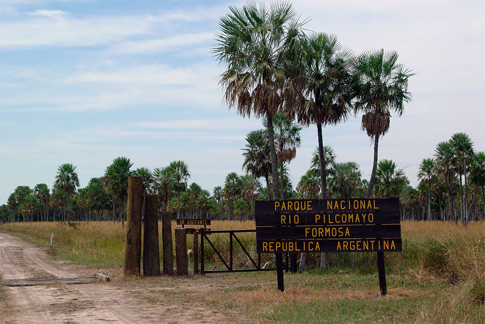 Dirt road leading to a gate with a sign that reads 'PARQUE NACIONAL RIO PILLCOMAYO FORMOSA REPUBLICA ARGENTINA', with palm trees and grassland in the background