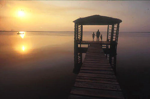 Wooden observation platform extending over calm water with three people silhouetted against sunset sky
