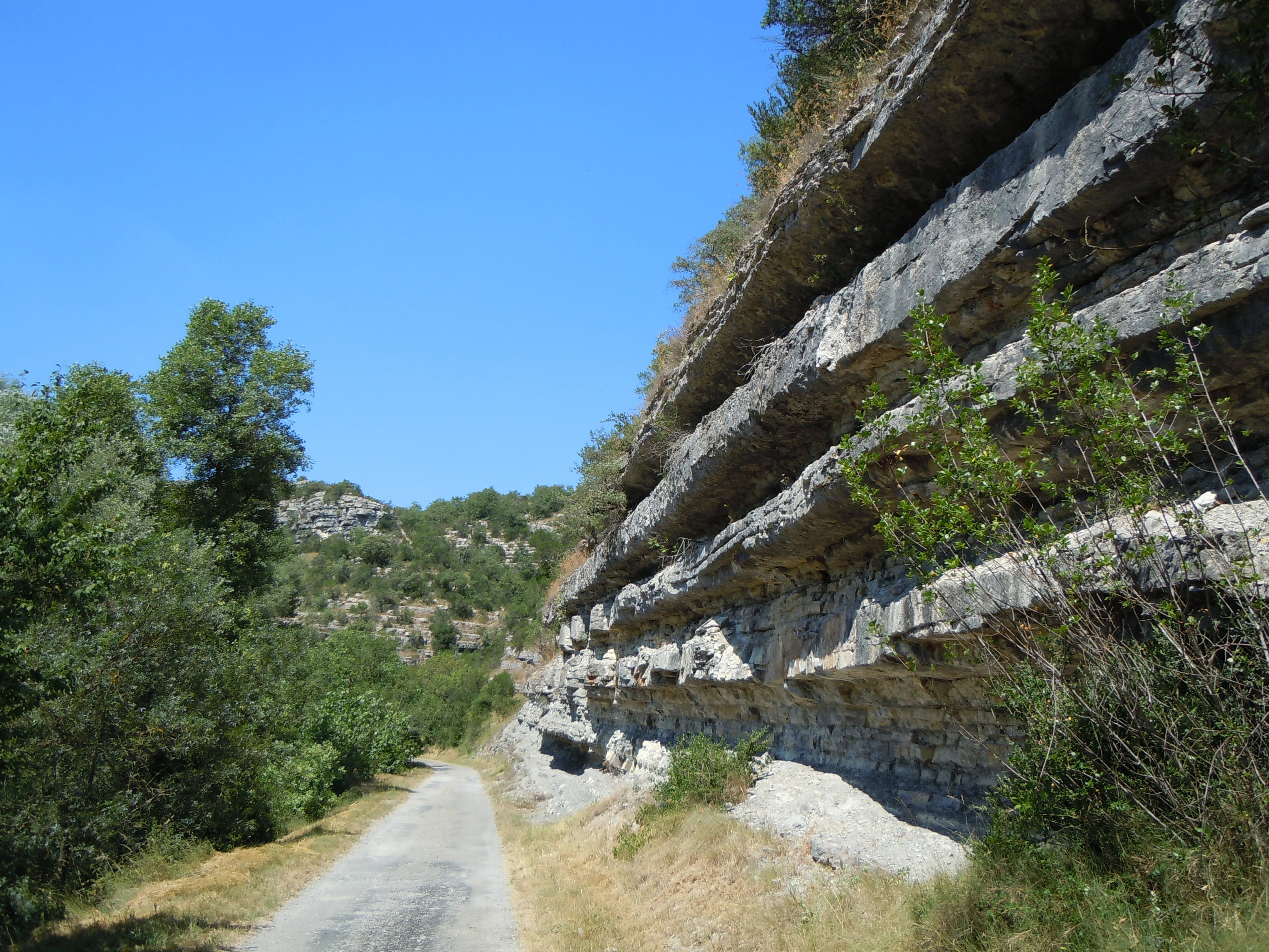 A gravel path runs alongside a large, layered limestone cliff face with green trees and vegetation on the left, under a clear blue sky.