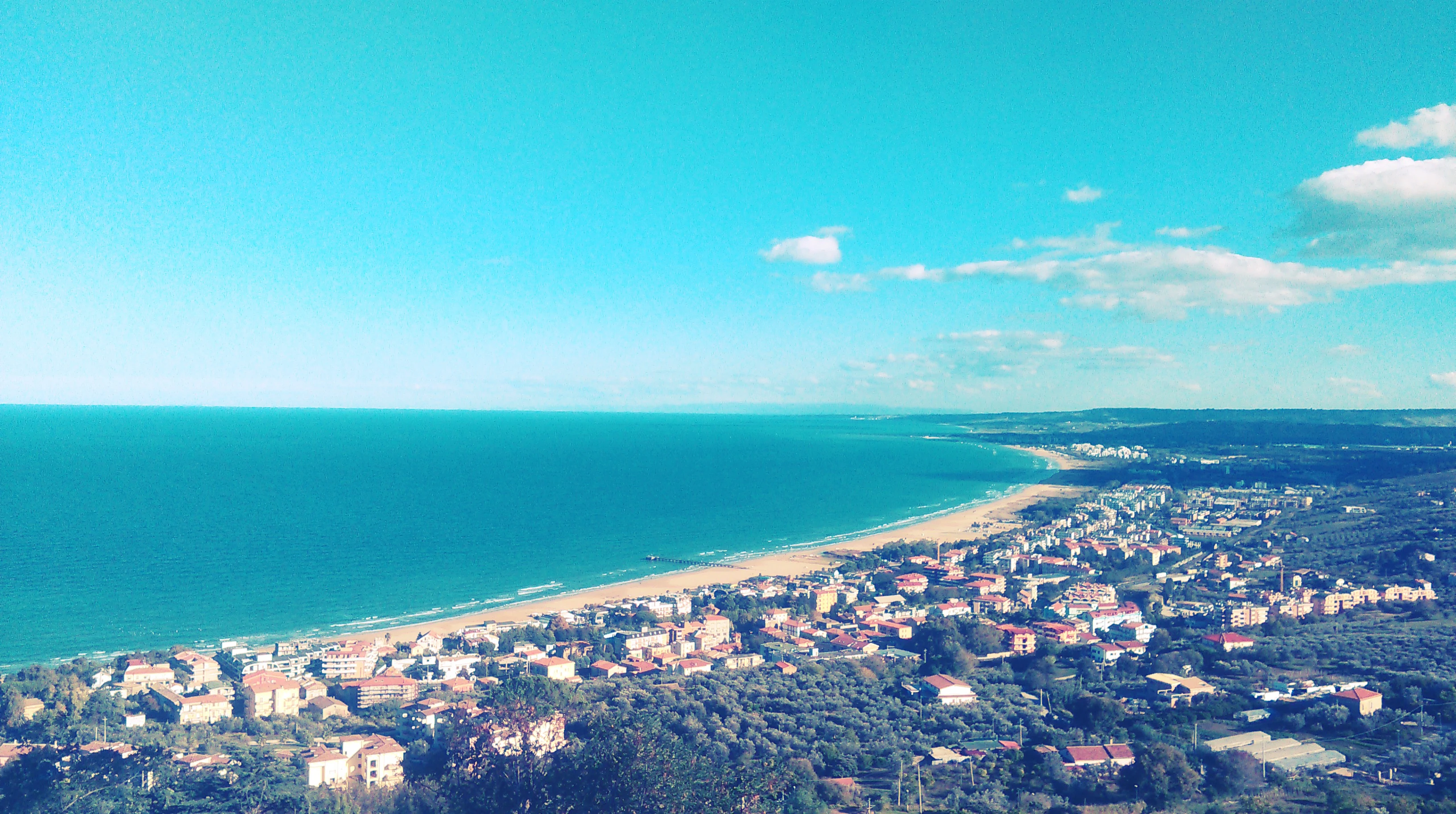 A wide landscape view of a coastal town along a long sandy beach next to a large body of blue water under a bright blue sky with scattered clouds.